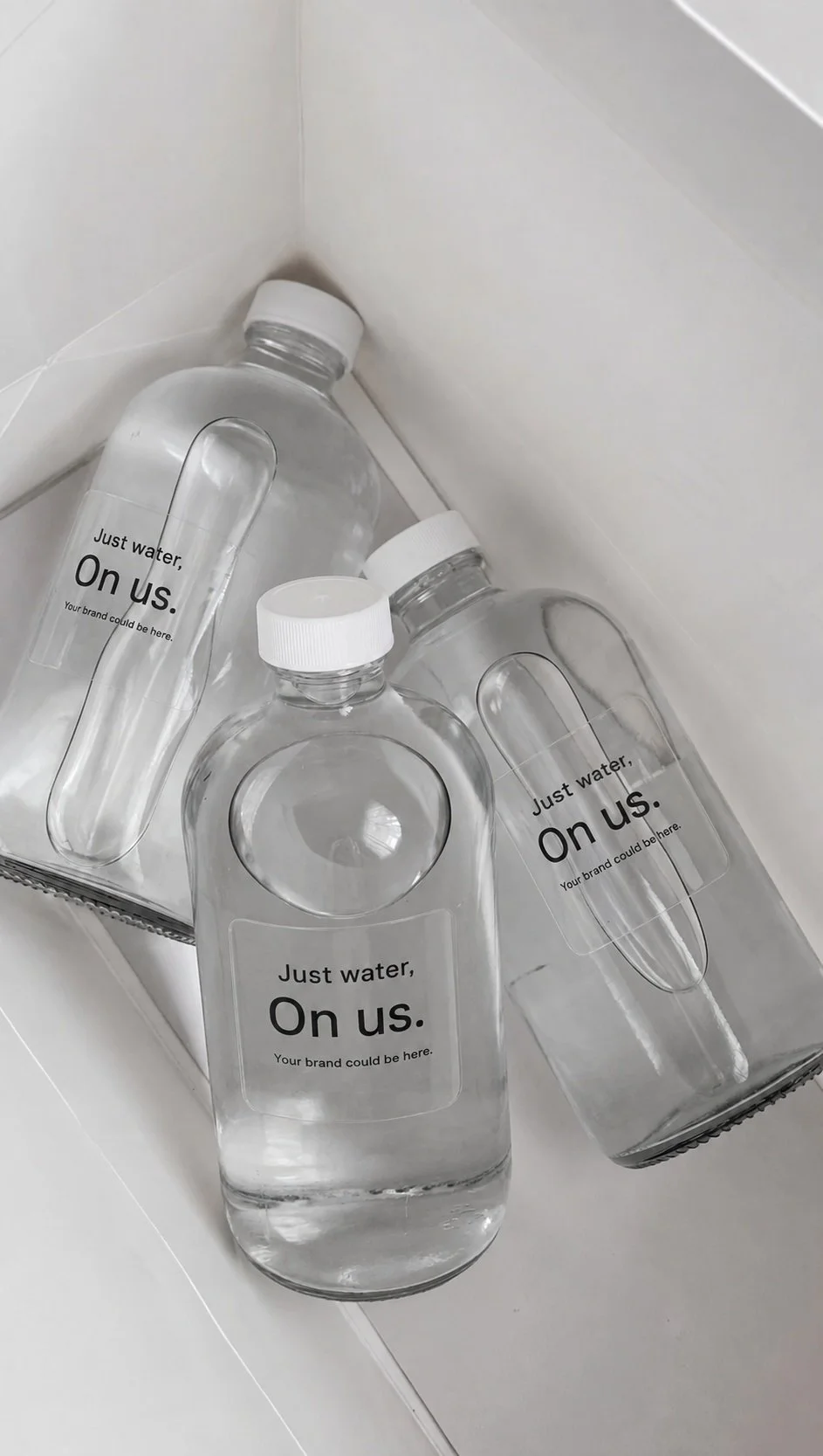 Several empty clear plastic water bottles with white caps, labeled "Just water, On us. Your brand could be here," placed in a white box on a white surface.