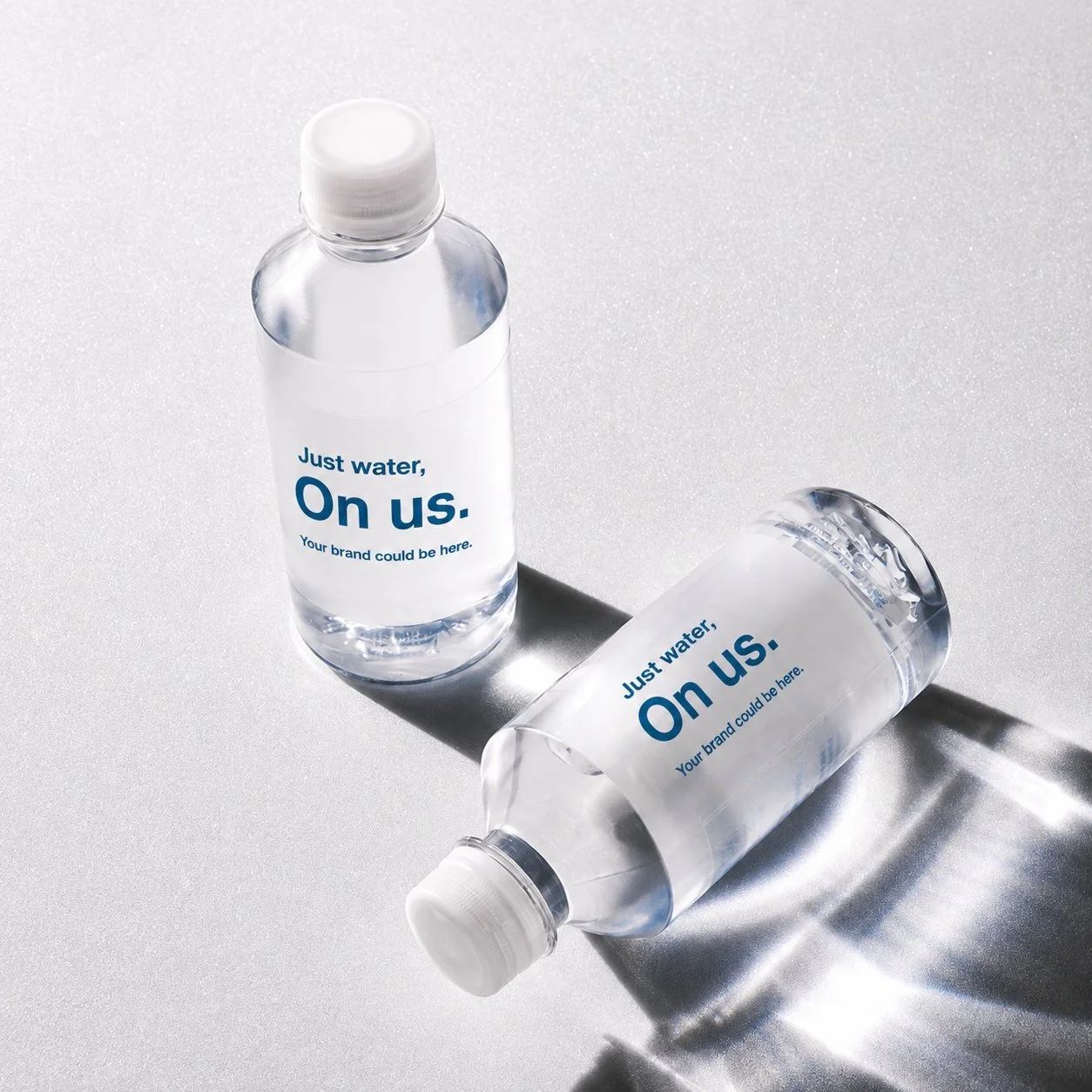 Two transparent water bottles with white caps, labeled "Just water, On us. Your brand could be here," placed on a metallic surface, casting shadows.