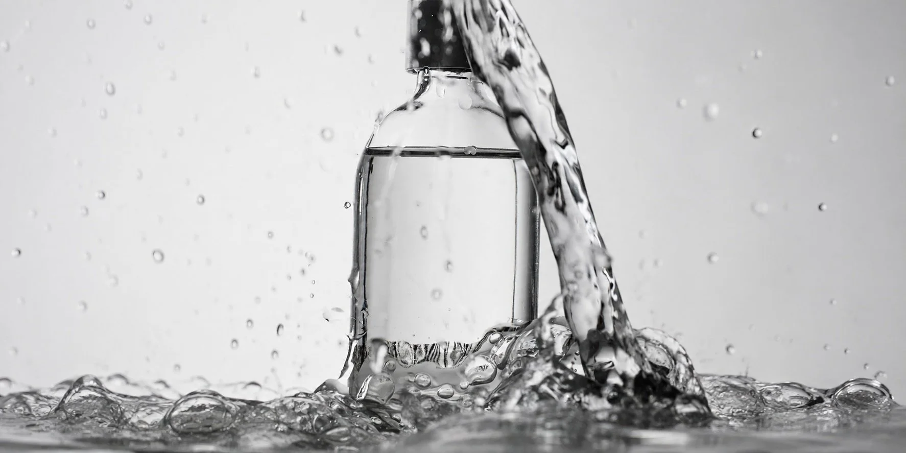 A close-up of a glass water bottle under a water faucet, with water splashing around as it flows into the bottle, captured in black and white.