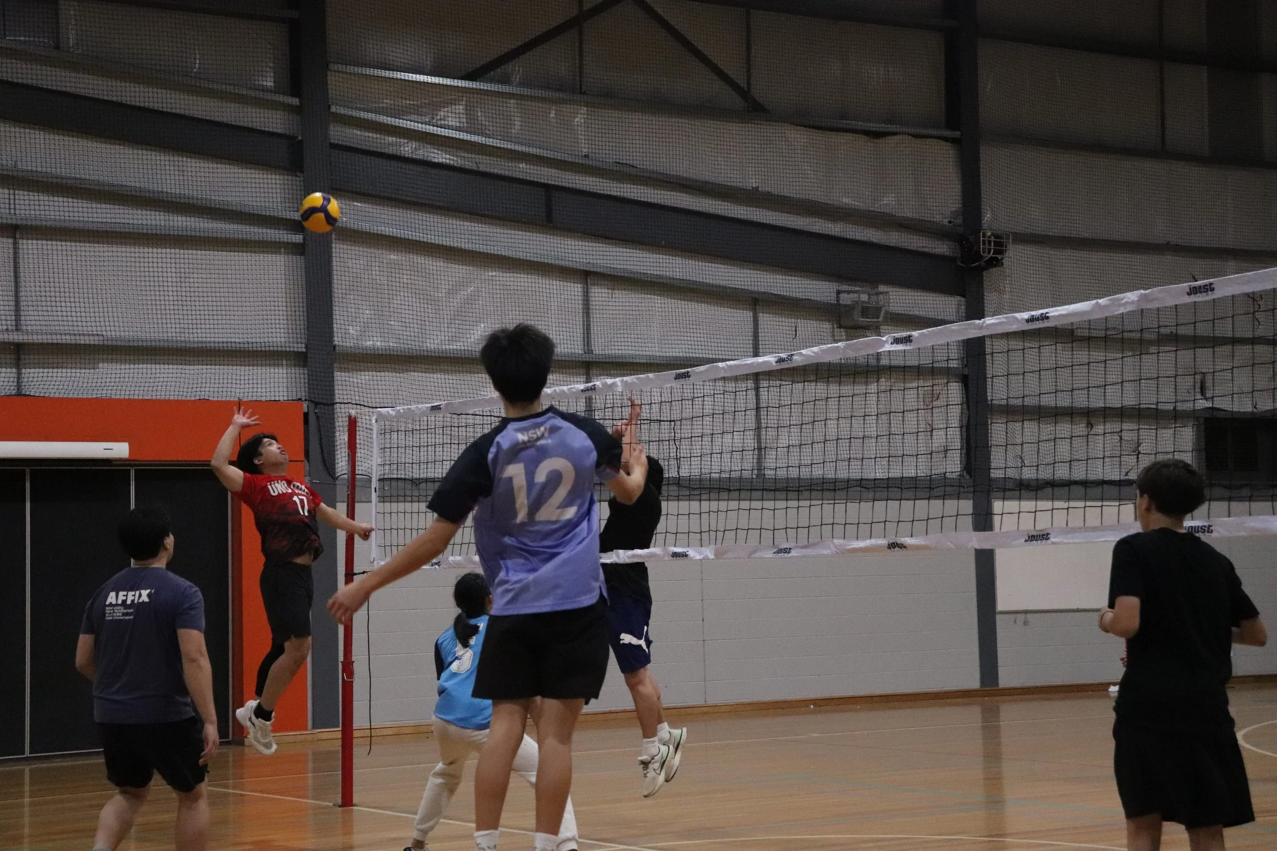 Indoor volleyball game with players, one jumping to hit the ball over the net, in a gymnasium.