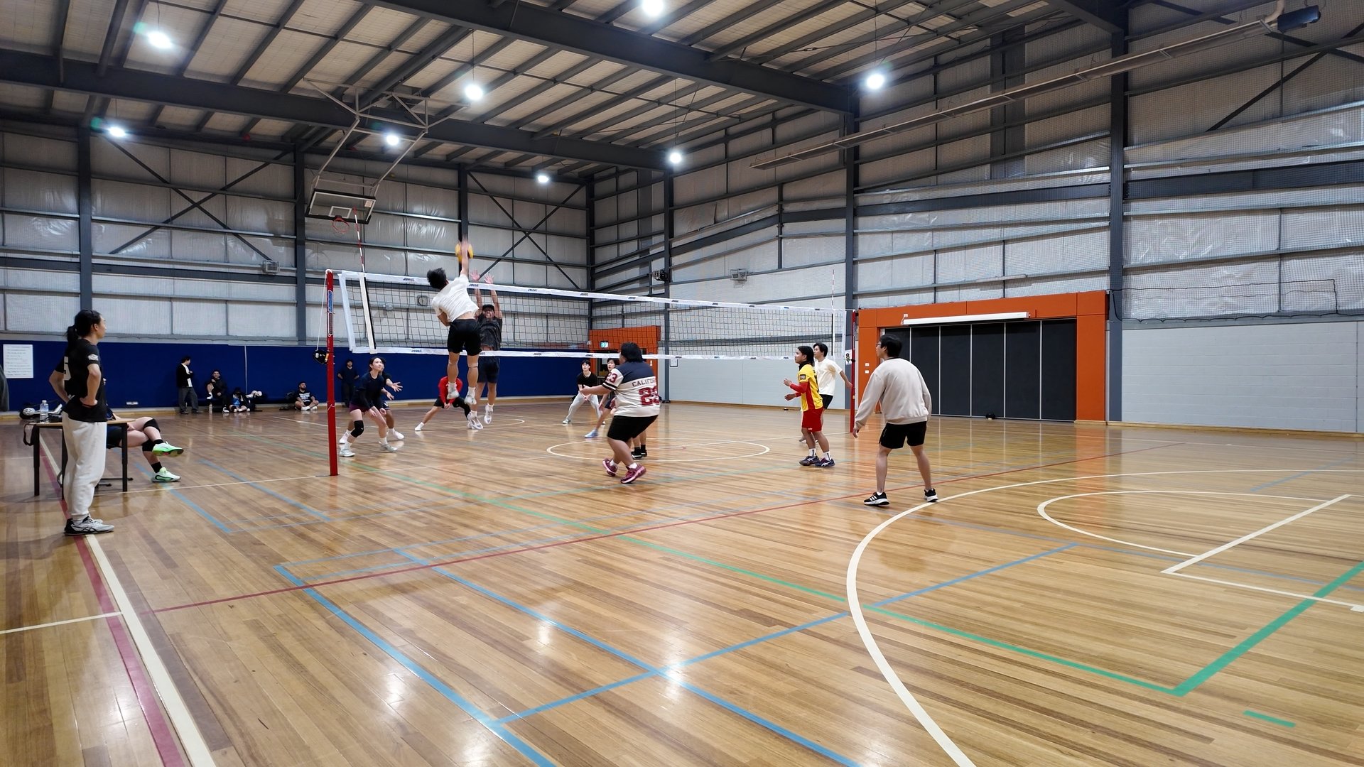 Indoor volleyball game with players jumping and preparing to hit the ball at the net, spectators on benches, and a referee on the sideline in a gymnasium.