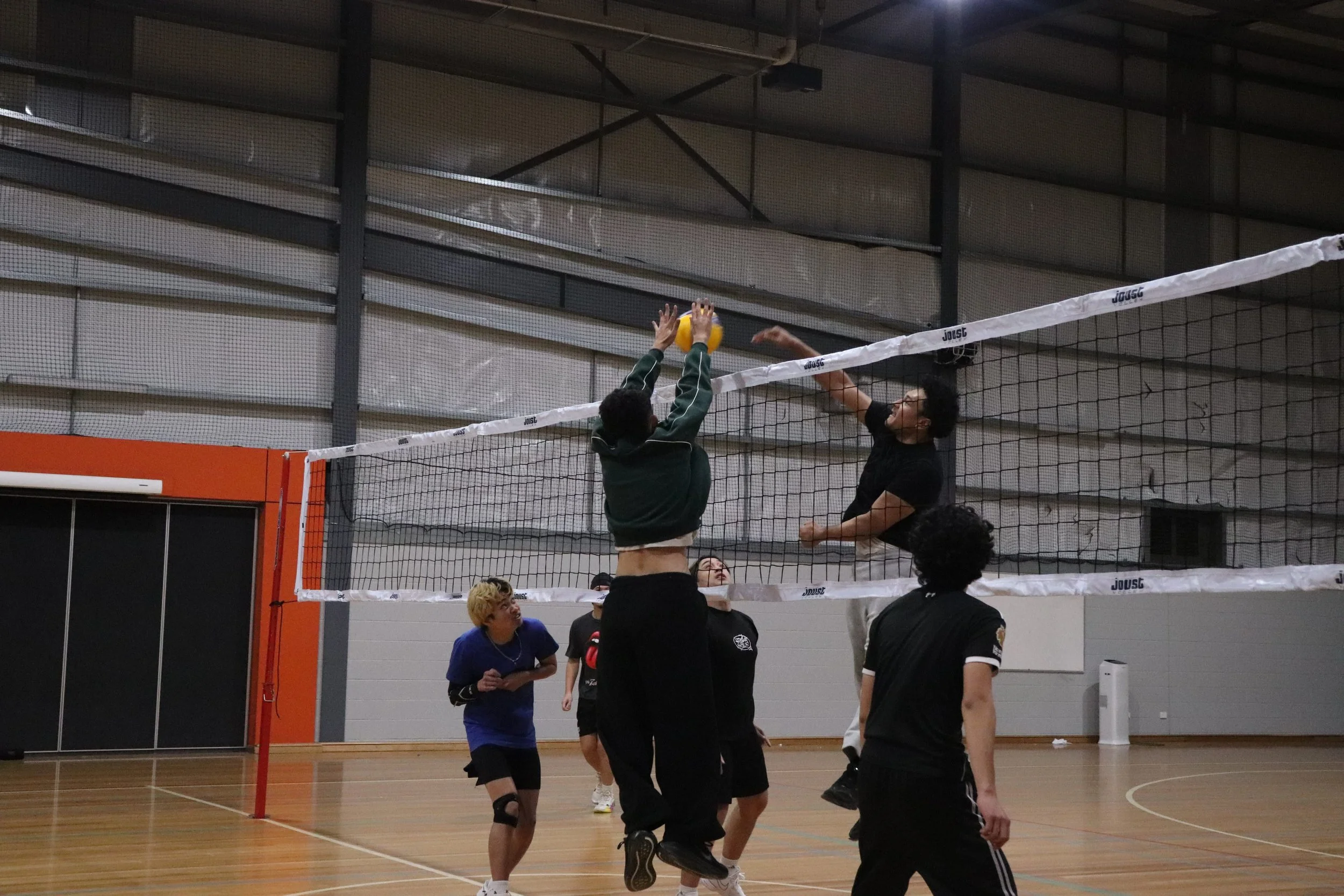 Two volleyball players are jumping at the net, one attempting to hit the ball while the other blocks. Several people are watching from below in an indoor gym with a high ceiling.