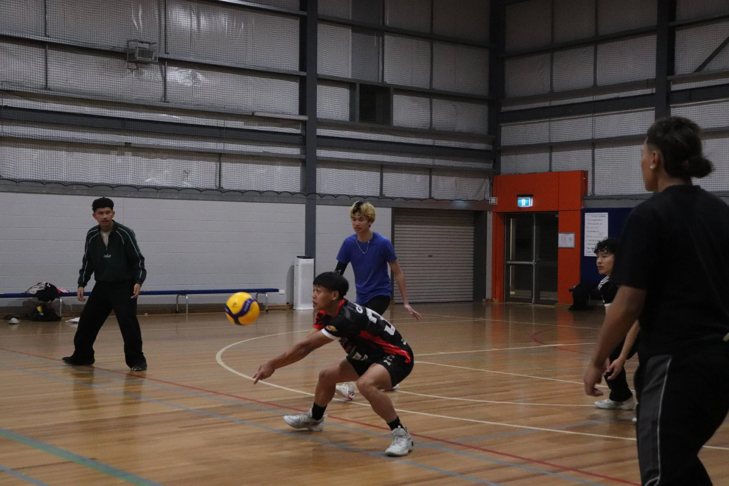 A group of young people playing indoor volleyball in a gymnasium. One player is diving to the ground attempting to hit the ball, while others watch and prepare to respond. The gym has a wooden floor, a large high ceiling, and a metal mesh wall.