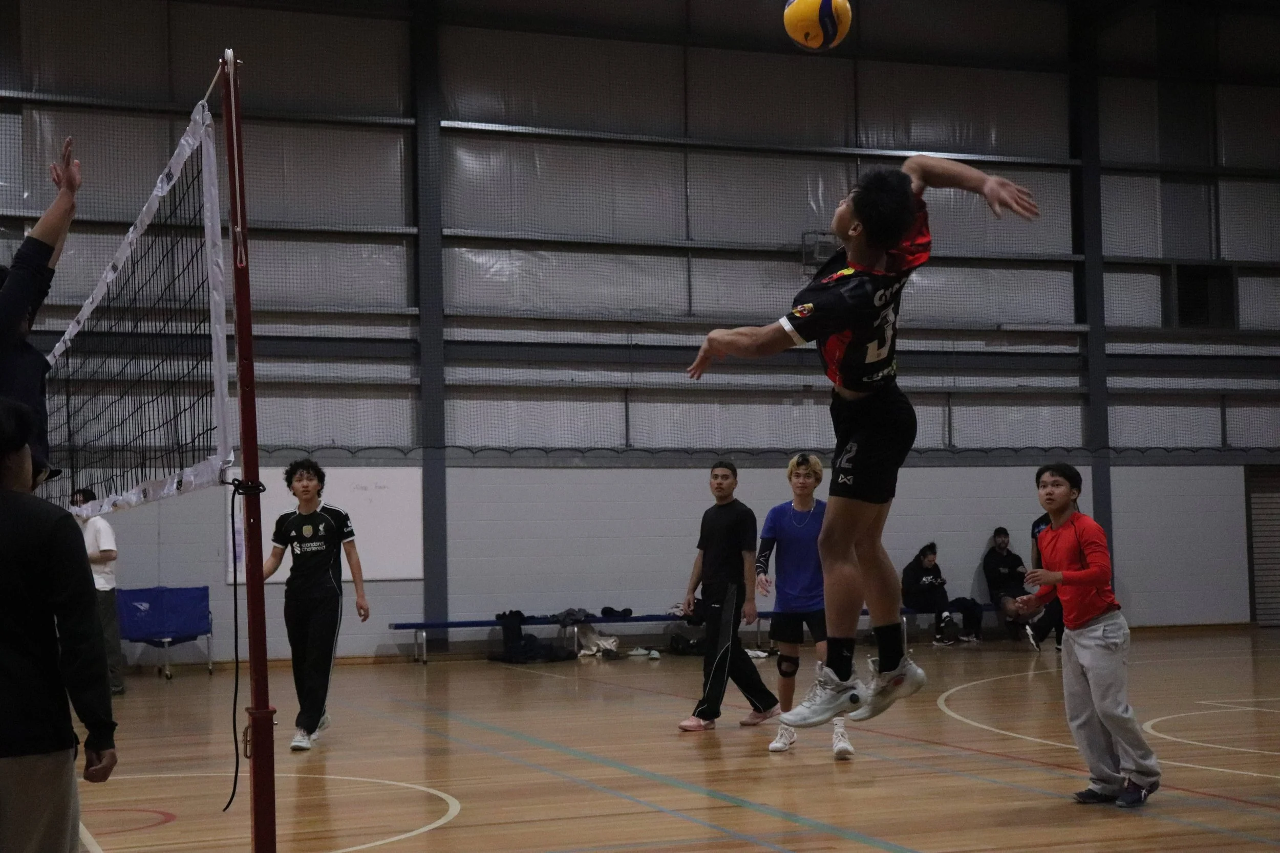 Indoor volleyball game in progress with players preparing to serve. One player in mid-air, others standing around court, spectators observing.