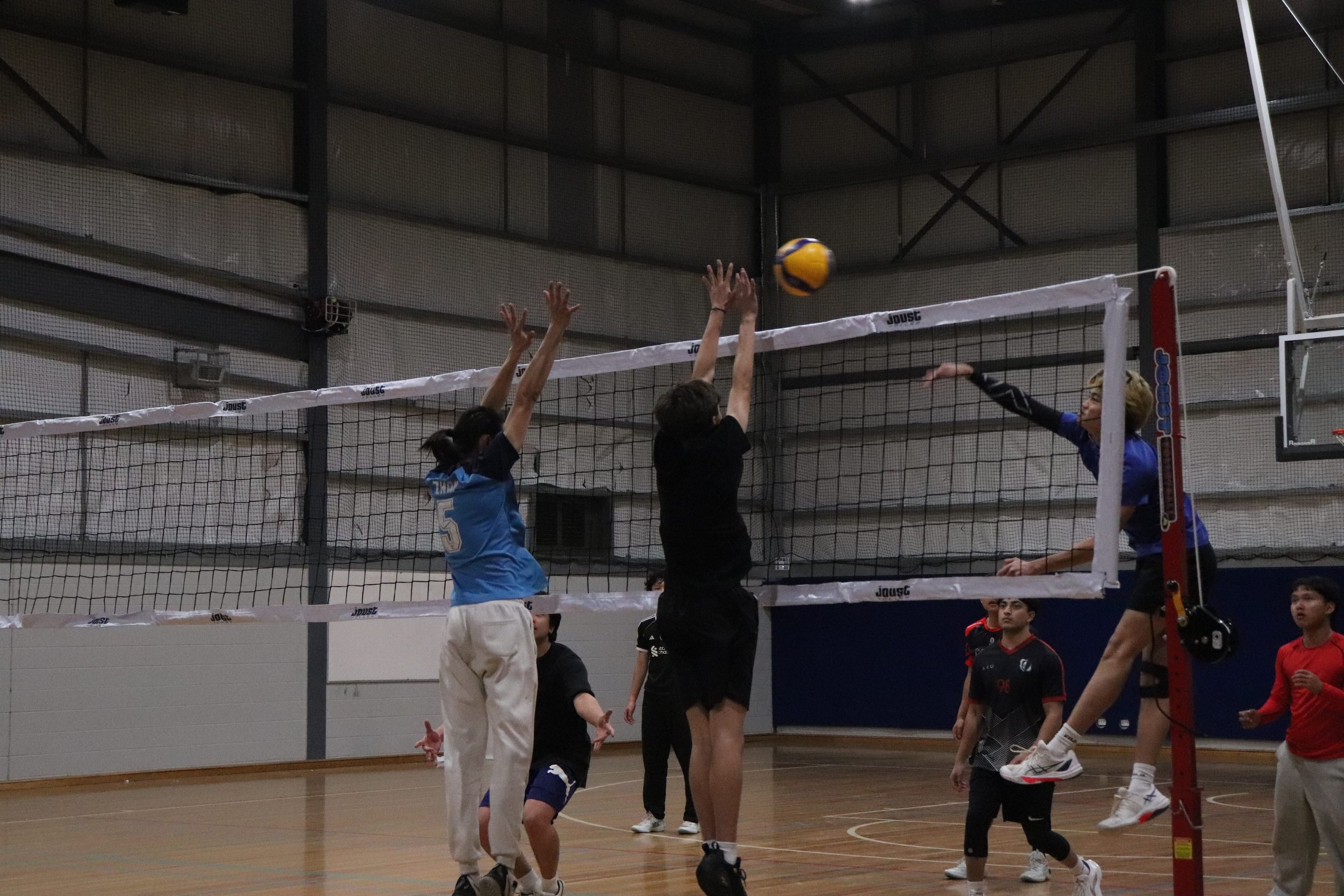 A volleyball game with players jumping to hit the ball over the net indoors.