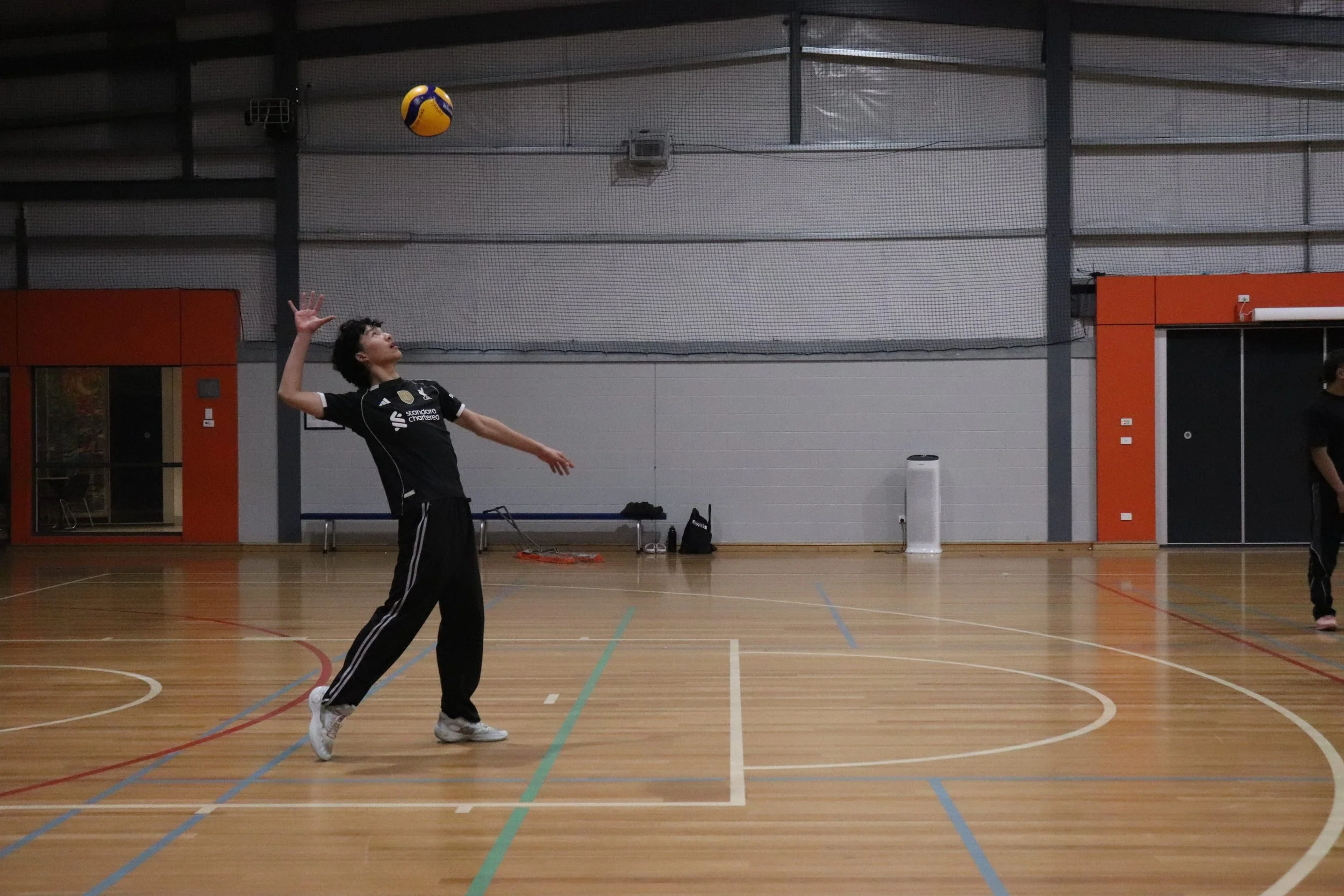 A young man playing volleyball indoors on a wooden court, preparing to serve the ball with a net visible in the background.