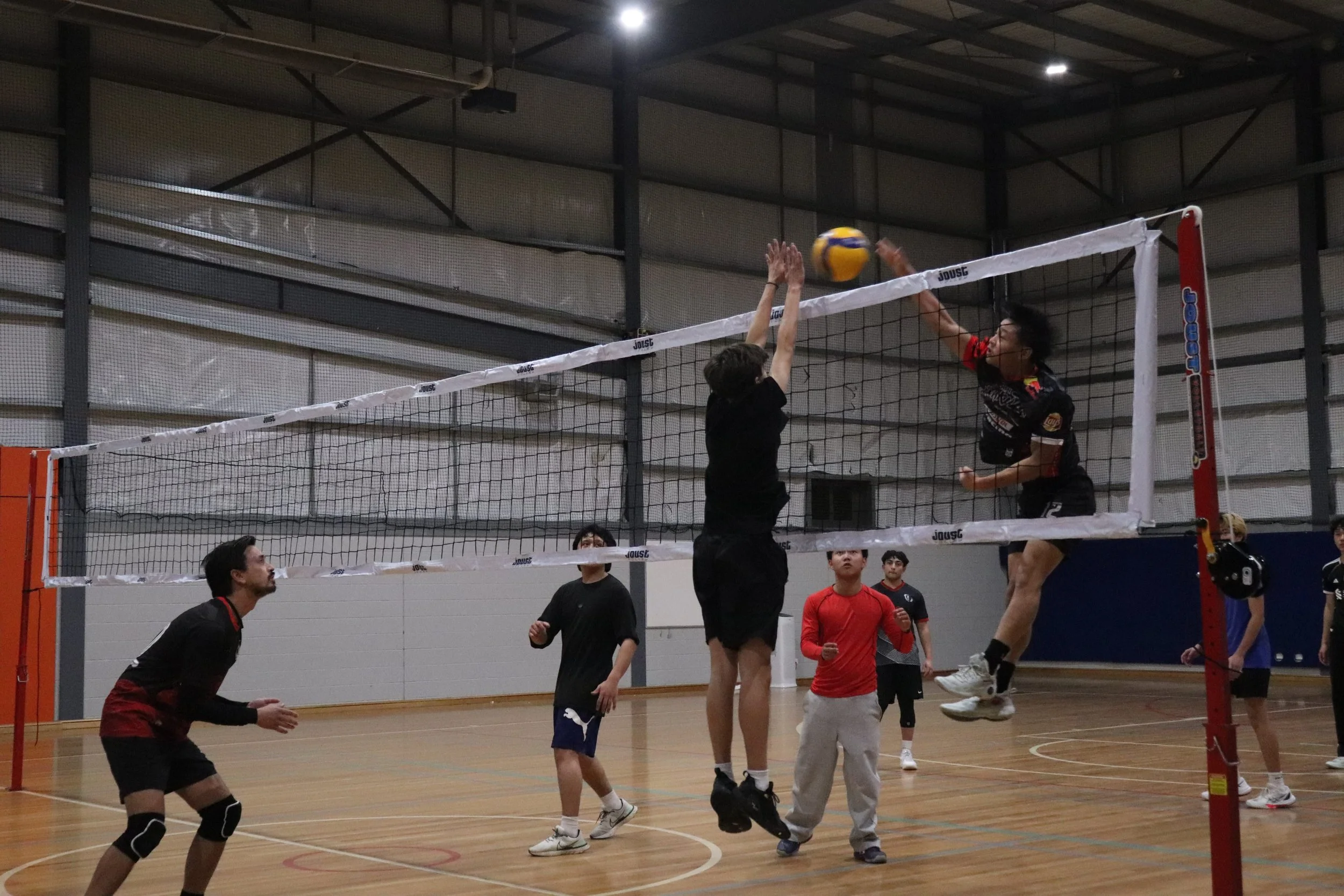 A group of people playing volleyball in an indoor gym. One person is jumping to hit the ball over the net, while others are watching and preparing to react.