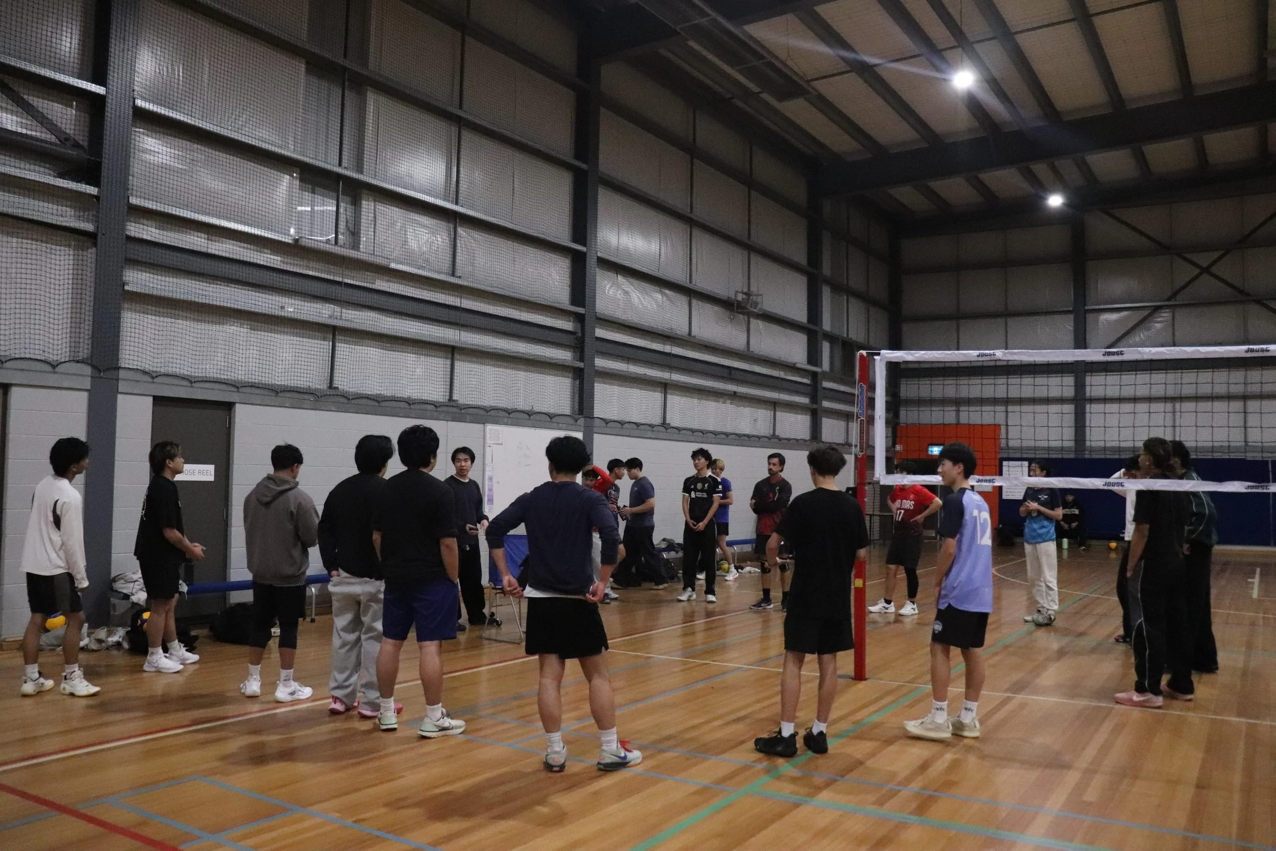 Indoor volleyball court with a group of people, some standing in line, others gathered around, and players preparing for a game. The court has a high ceiling, metal walls, and a volleyball net. Overhead lights illuminate the space.