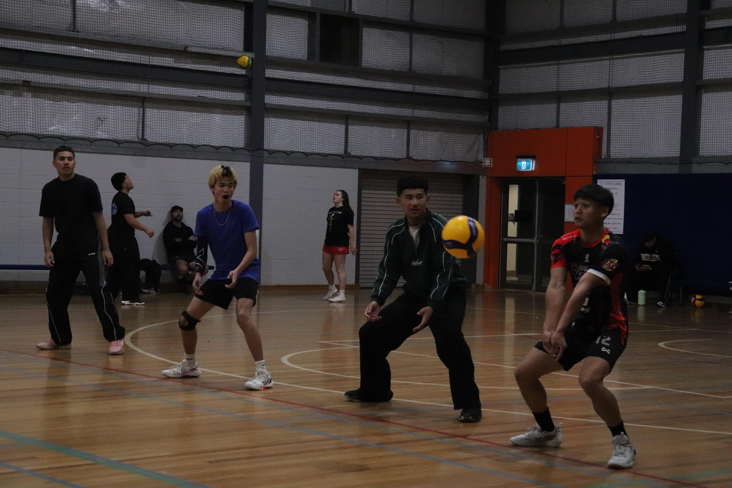 A group of young people playing indoor volleyball in a gymnasium. Several players are actively engaged, with one preparing to hit the ball. The gym has a wooden floor, high ceilings, and netting along the walls.