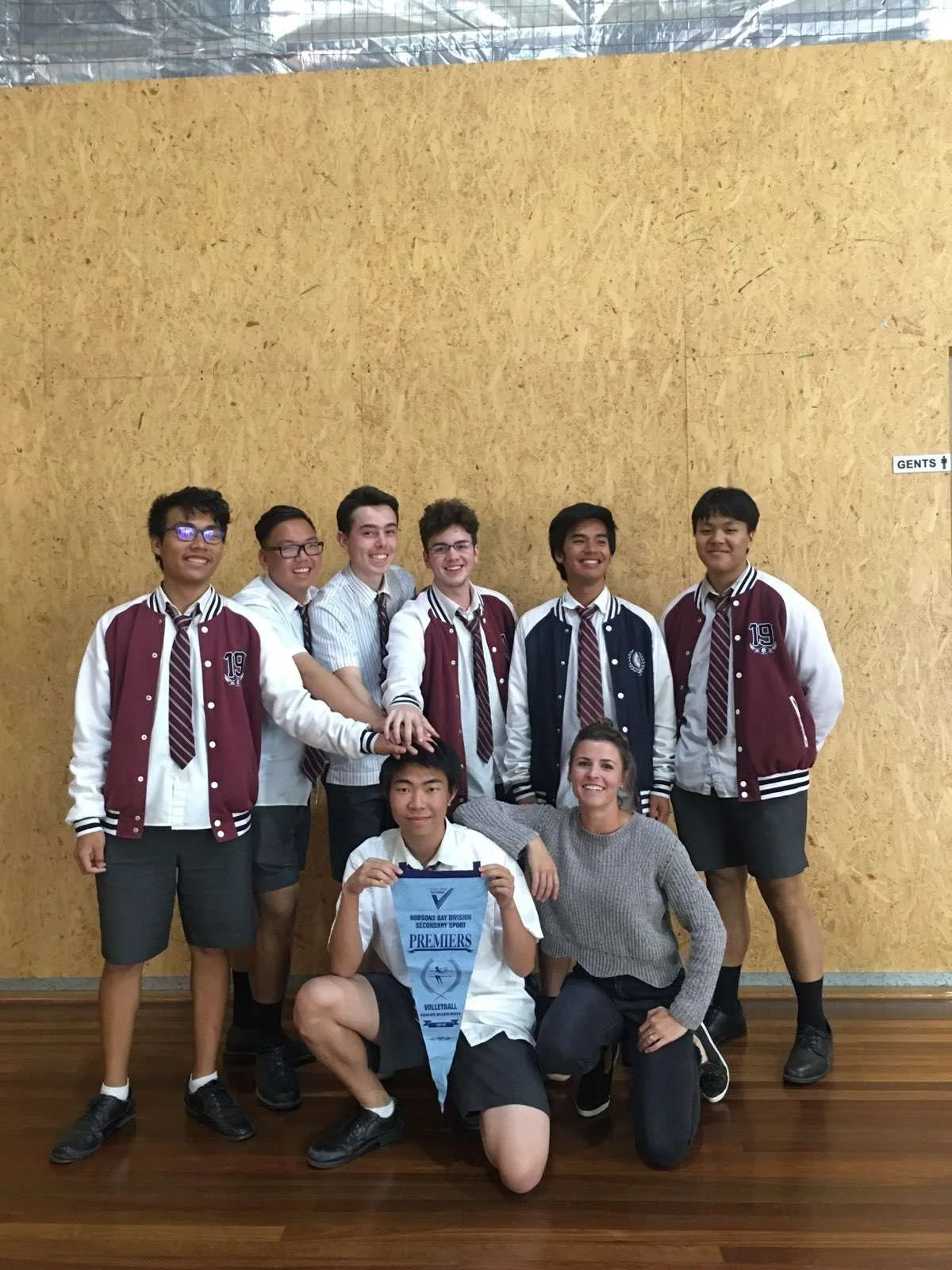 A group of students and a woman posing in a gym, with one student holding a volleyball banner. The students are wearing school uniforms, some with varsity jackets, and everyone is smiling.