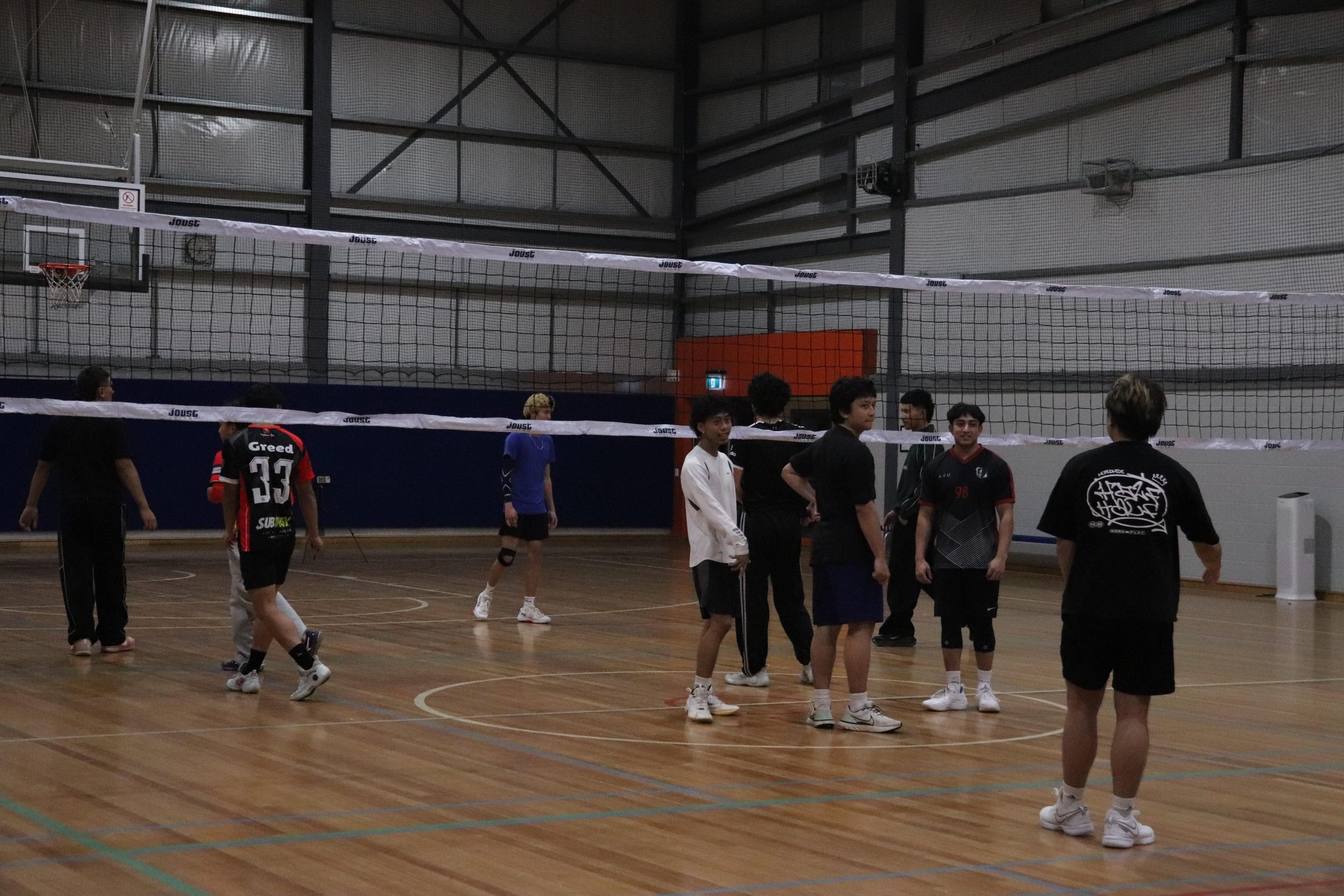 A group of young men in sportswear gathered inside a gymnasium with wooden floors, a volleyball net, and basketball hoops, preparing for or engaging in a volleyball game.