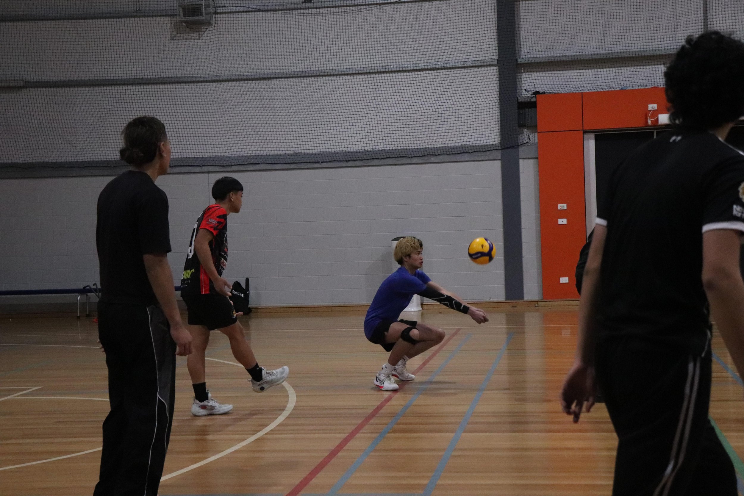 A group of people playing volleyball indoors on a wooden court with a net, with some players in motion preparing to hit or receive the ball.