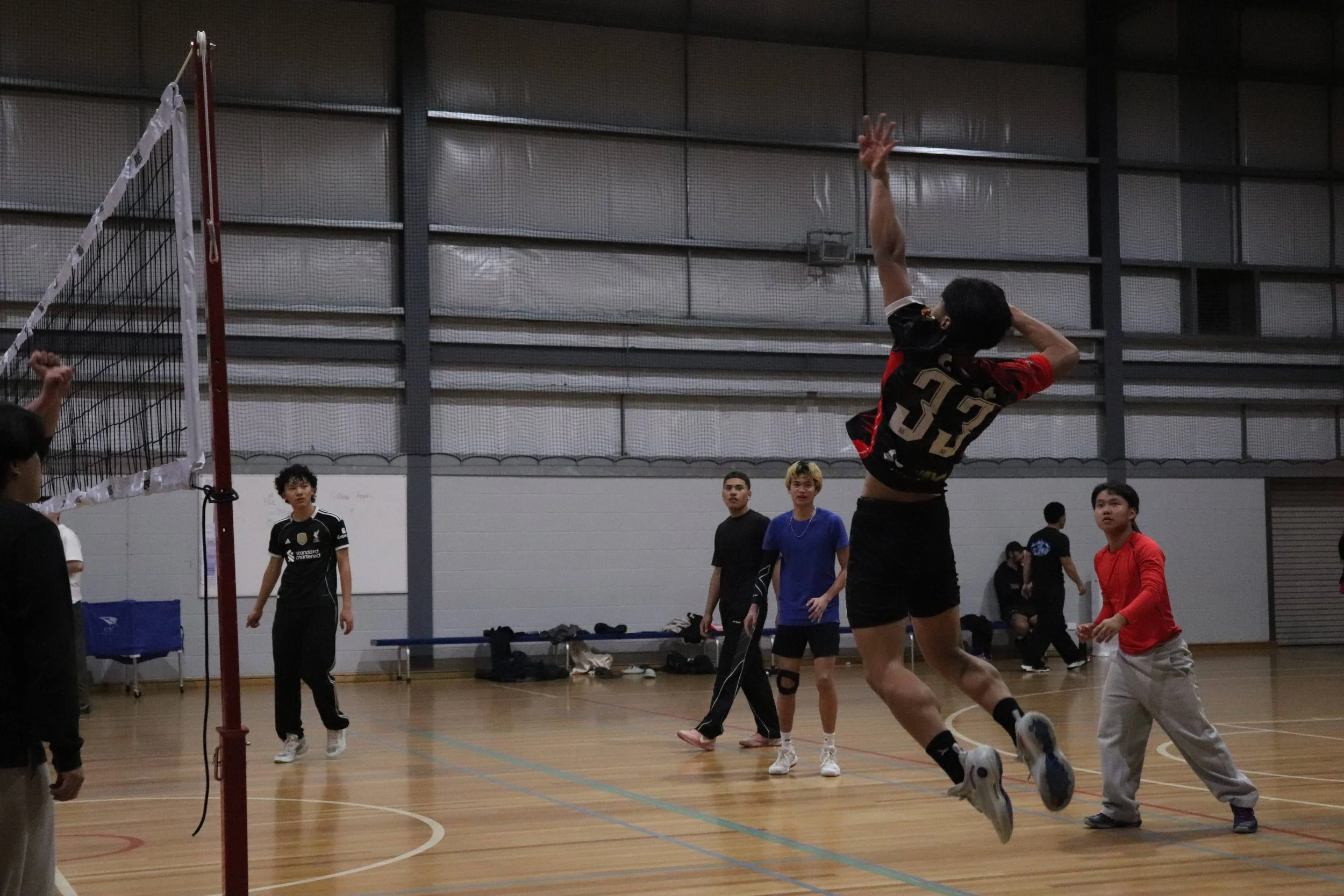 A group of young men playing volleyball indoors, one jumping to hit the ball over the net, while others watch and prepare to play.