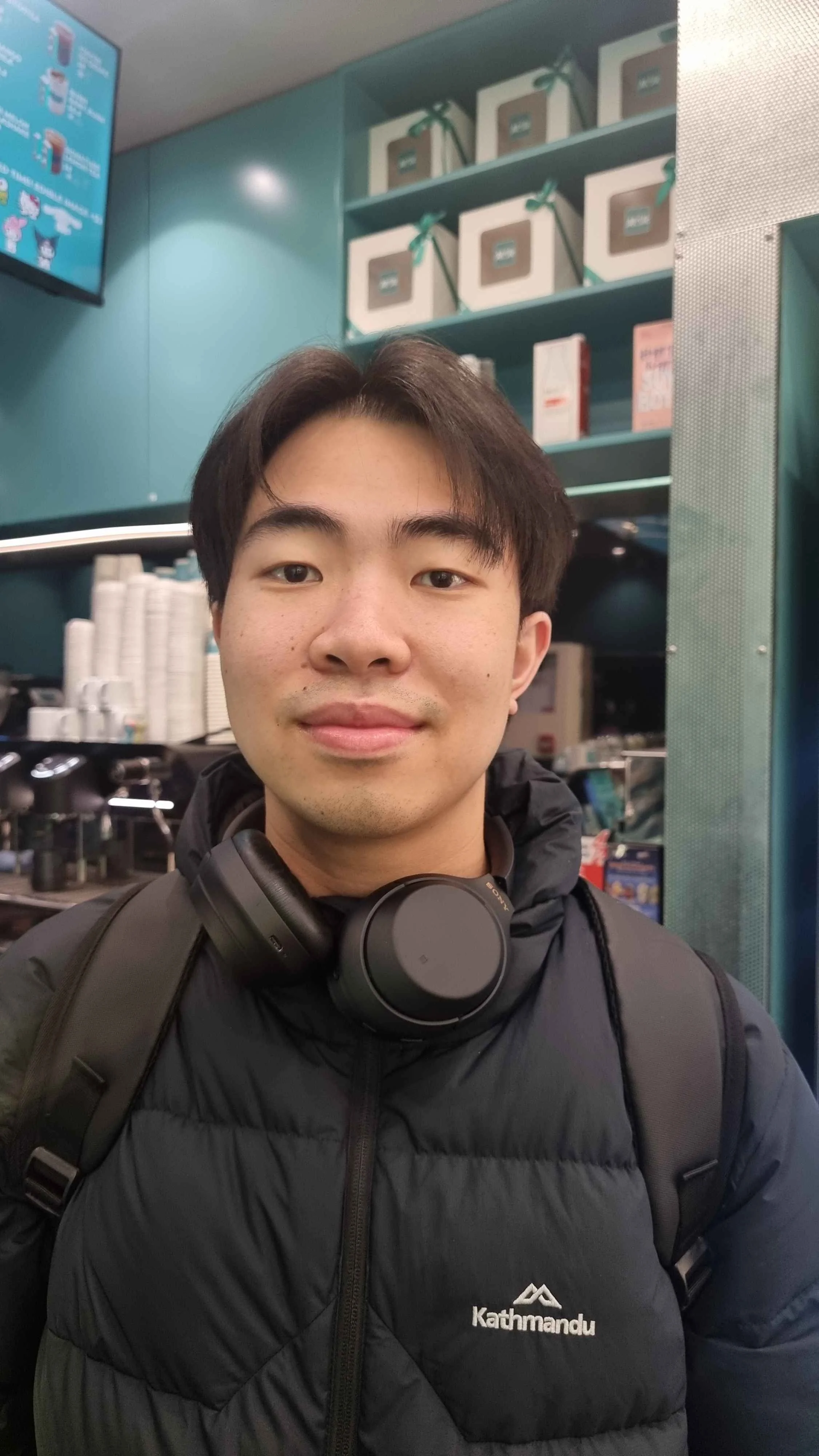Young man with black hair, wearing a black Kathmandu jacket with a backpack and headphones around his neck, standing inside a store with shelves of cups and boxes in the background.