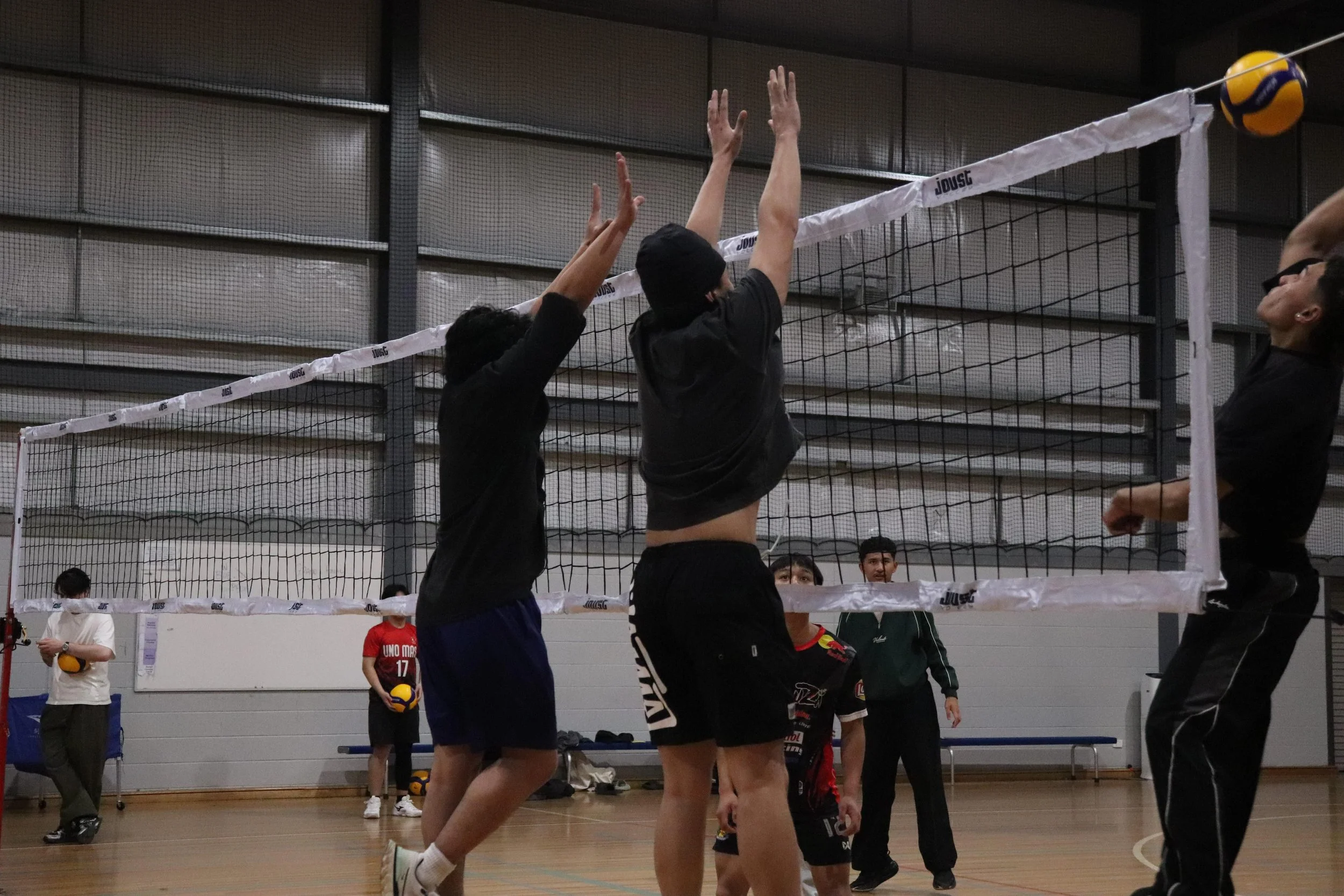 Indoor volleyball game with players jumping near the net, attempting to hit the ball. Spectators and other players are visible in the background.