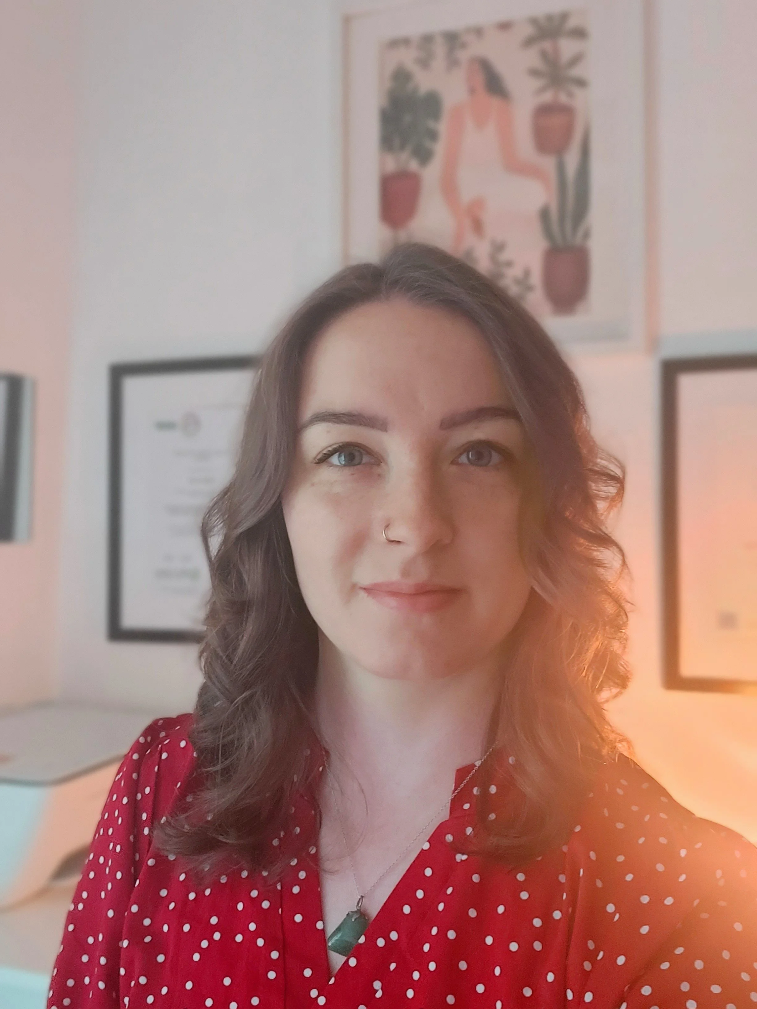 A young woman with curly brown hair wearing a red dress with white polka dots, standing indoors with framed artwork and certificates on the wall behind her.