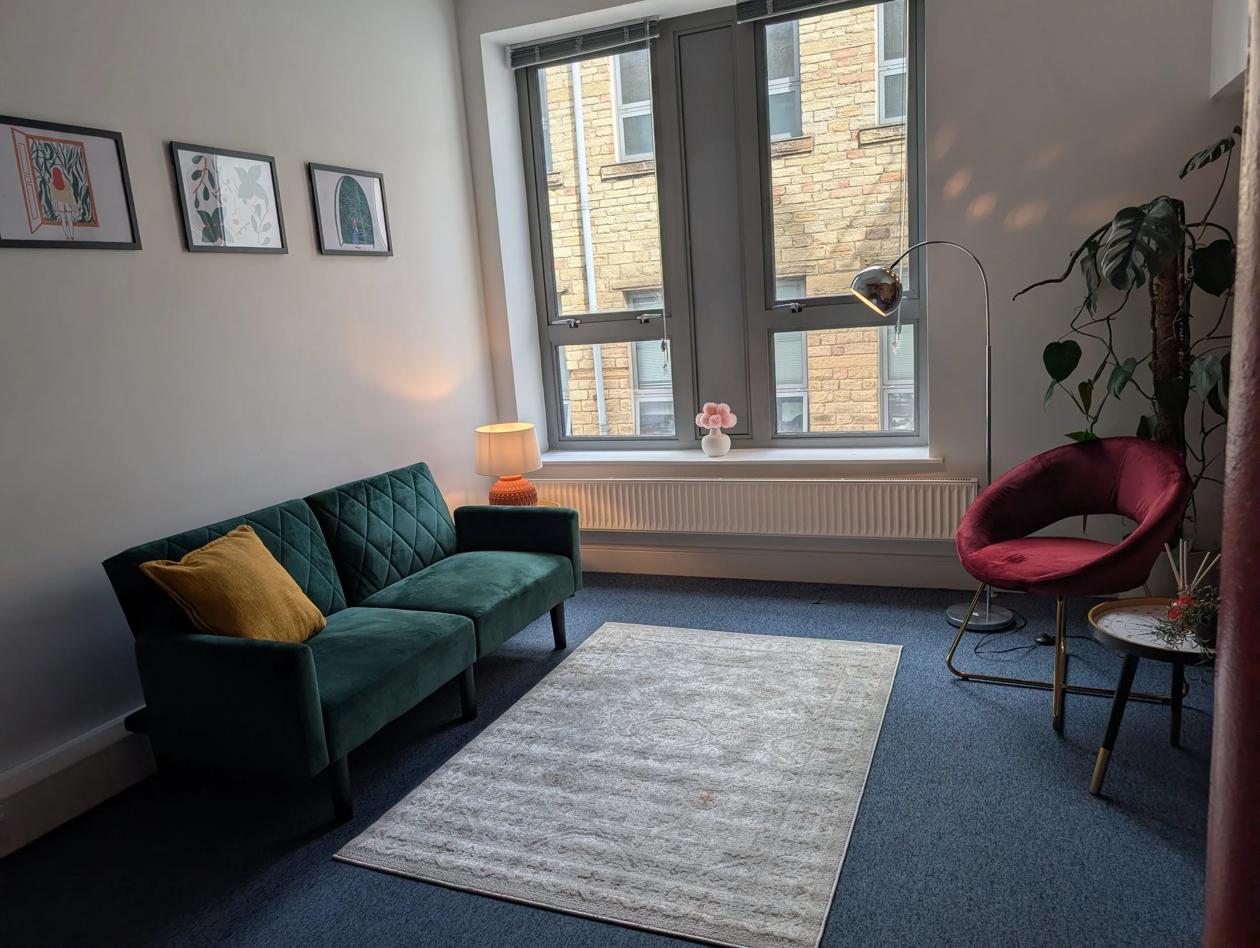 A cozy living room with a green velvet sofa and mustard yellow pillow, a pink armchair, a small side table with a flower arrangement, a desk lamp, and framed art on the wall. Large windows reveal a brick building outside.