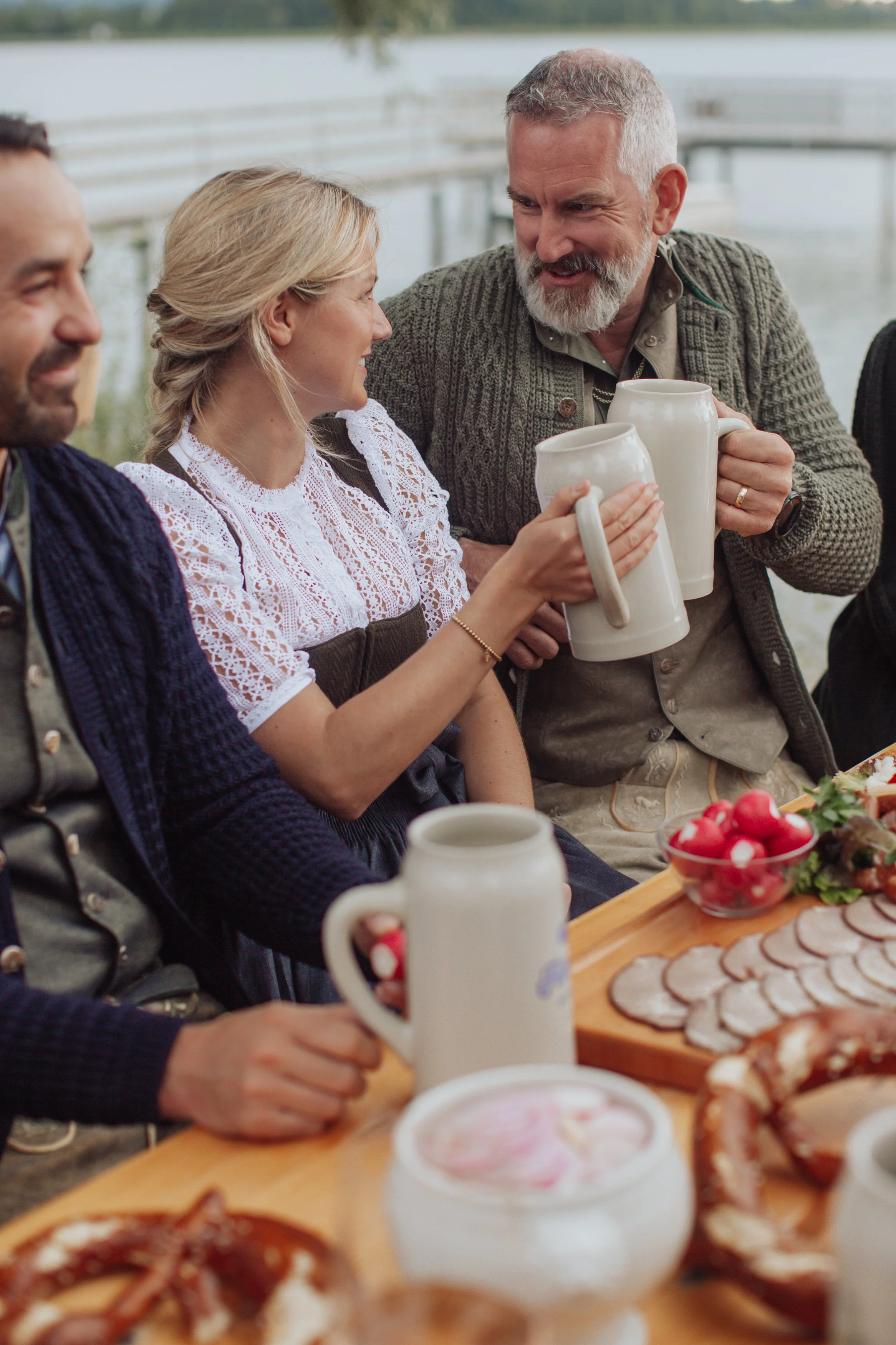 Gruppen von Menschen bei einem gemütlichen Mahl im Freien, mit Kaffee, Fladenbrot, Aufschnitt, Tomaten und Zwiebeln auf einem Holztisch, im Hintergrund einer Bademöglichkeit mit Wasser und Steg.