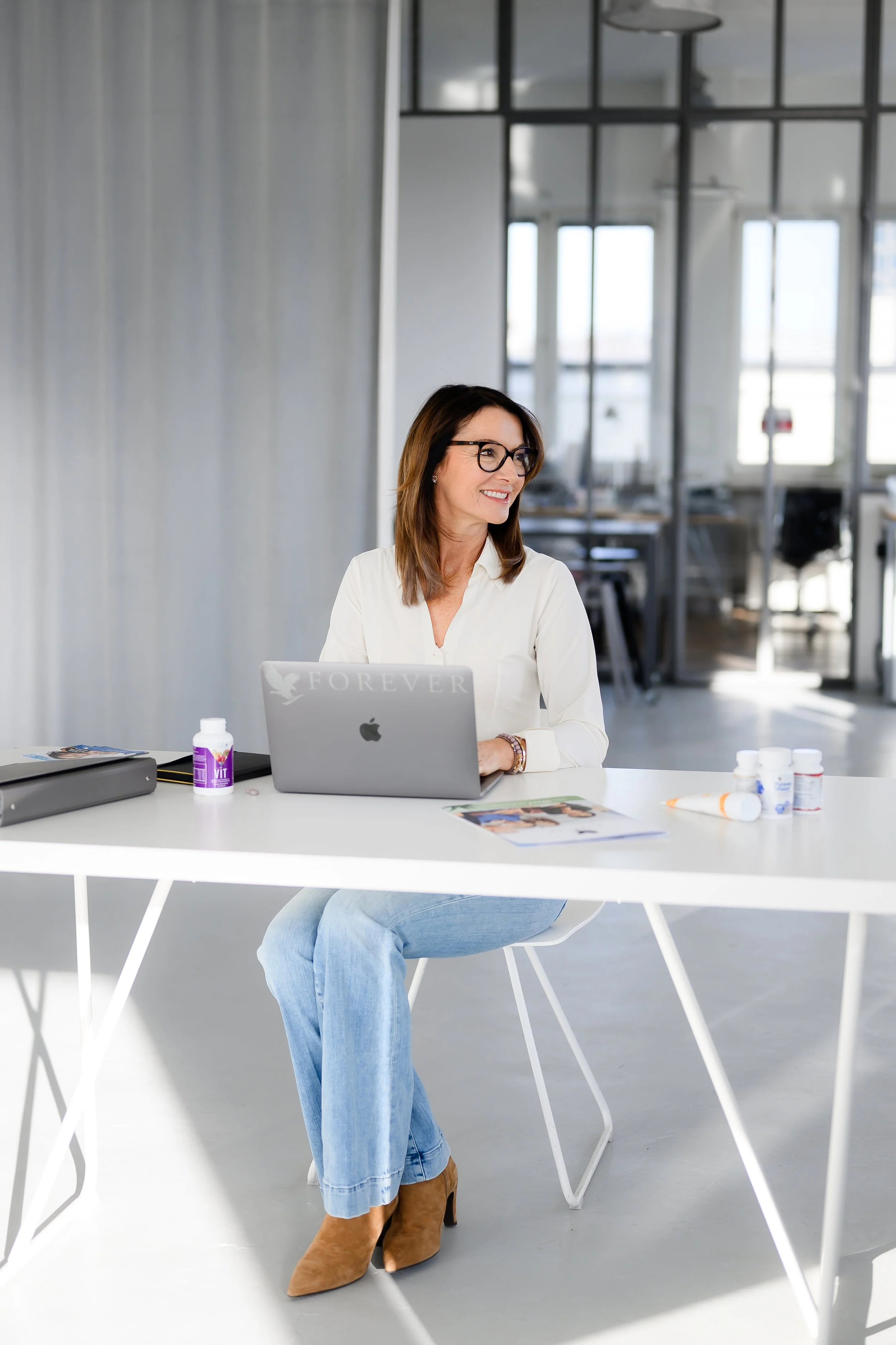 Frau mit Brille sitzt an einem weißen Tisch in einem modernen Büro, trägt eine weiße Bluse, hellblaue Jeans, braune Stiefel und hat braune, schulterlange Haare.