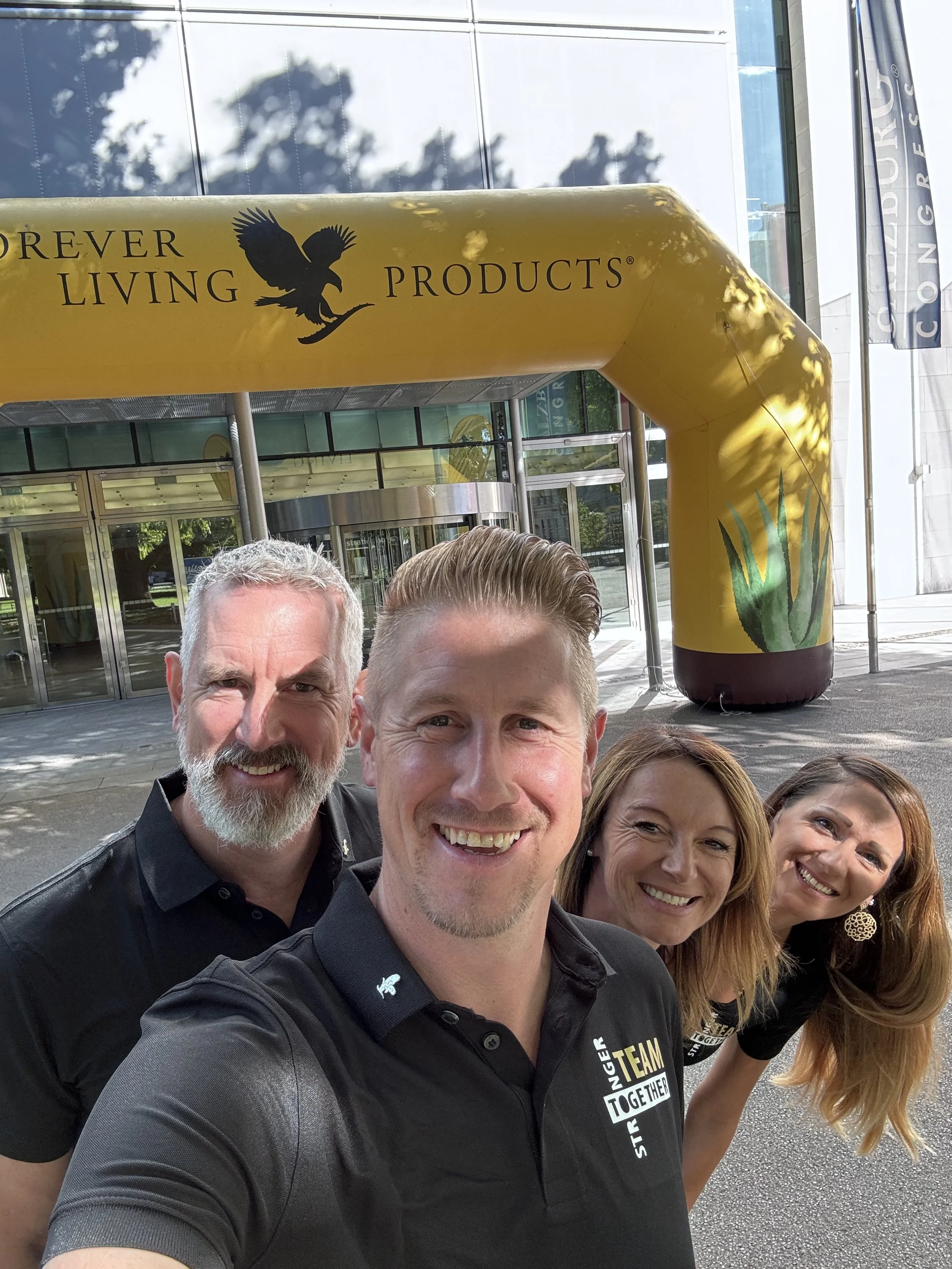 Gruppe von fünf Menschen beim Fotografieren vor dem Eingangsbereich der Messe, mit gelbem Luftballonbogen "Forever Living Products" im Hintergrund.
