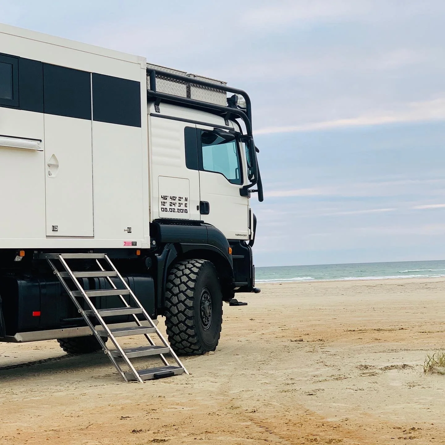 Ein geländegängiges Fahrzeug mit einem Aufbau steht am Strand, Blick auf das Meer und den Himmel im Hintergrund.