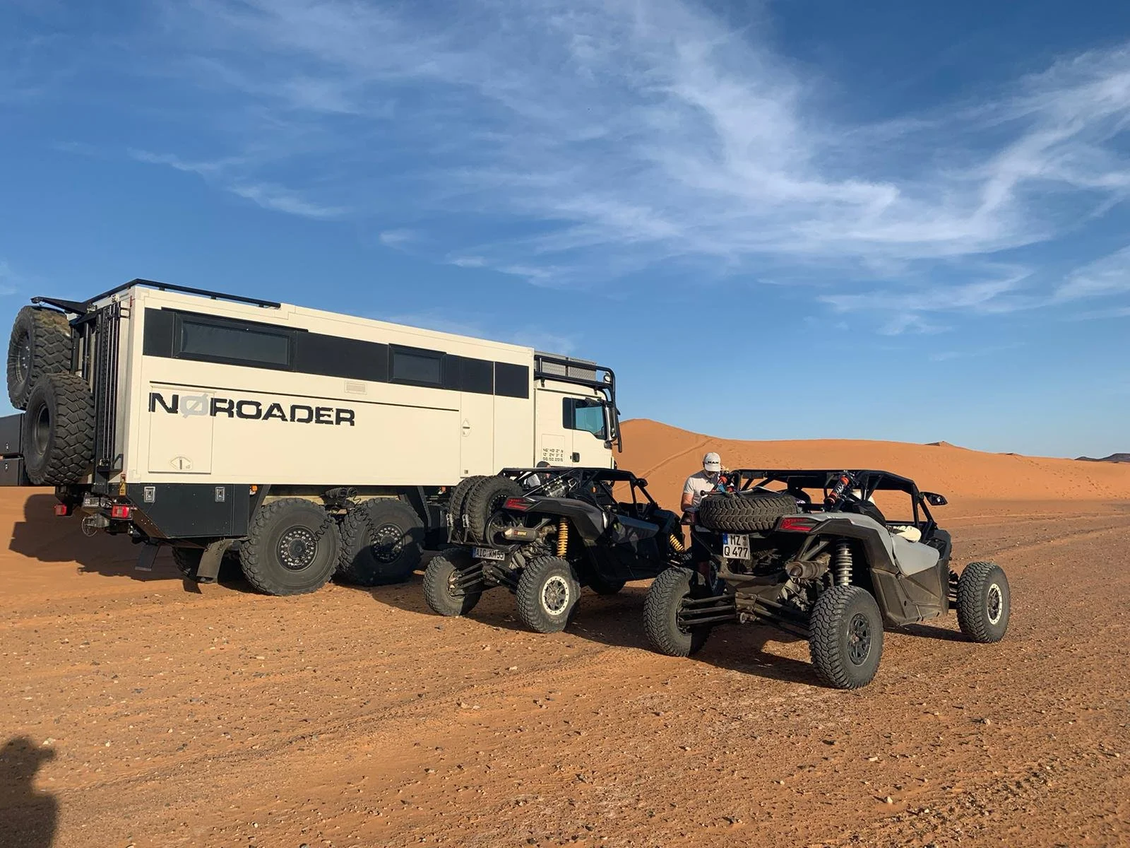 Ein offener Geländewagen und zwei kleinere Offroad-Fahrzeuge in einer Wüstenlandschaft mit Sanddünen, vor einem blauen Himmel. Es scheint sich um eine Expedition oder ein Abenteuer zu handeln.