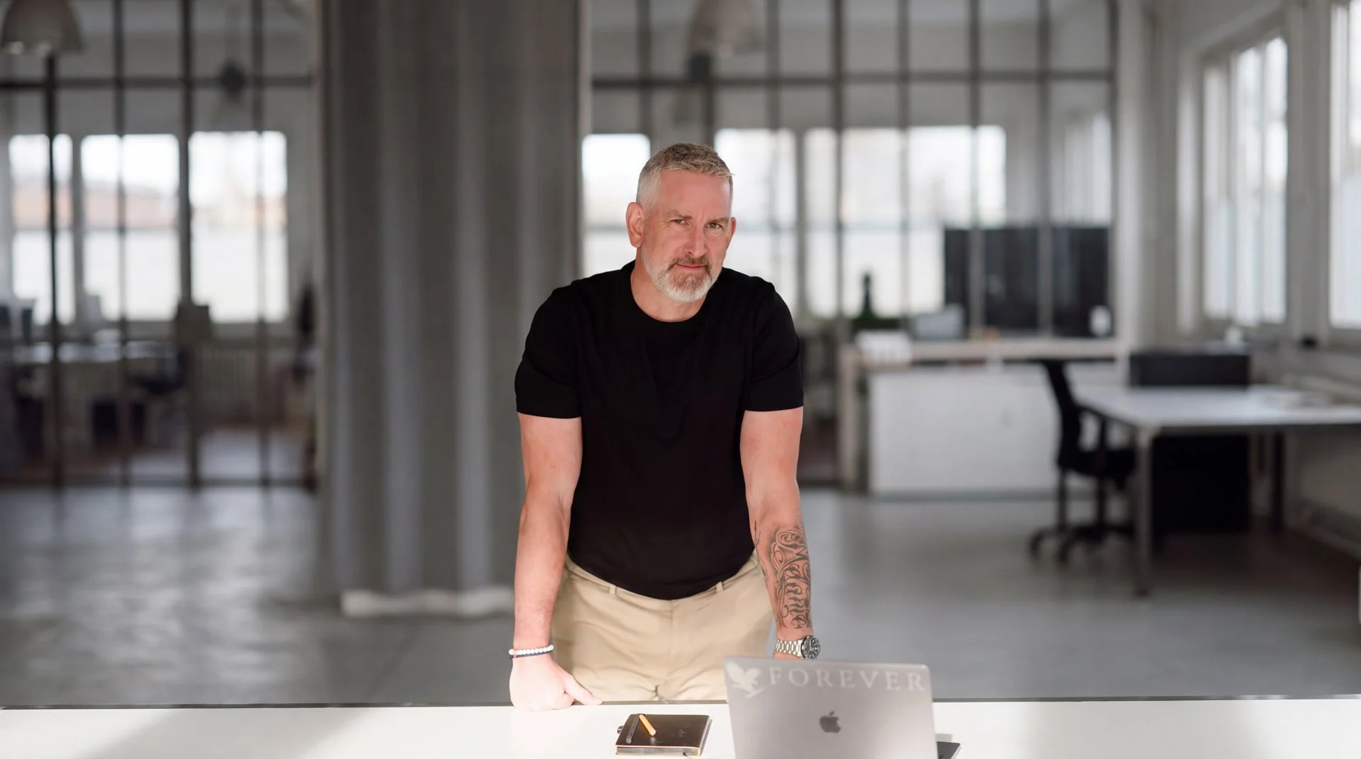 Ein Mann mit kurzen grauen Haaren und einem grauen Bart steht an einem weißen Tisch in einem modernen Büro, trägt ein schwarzes T-Shirt und khakifarbene Hosen, vor ihm auf dem Tisch liegen ein Notizbuch, ein Smartphone, ein Laptop und ein Computerbildschirm.