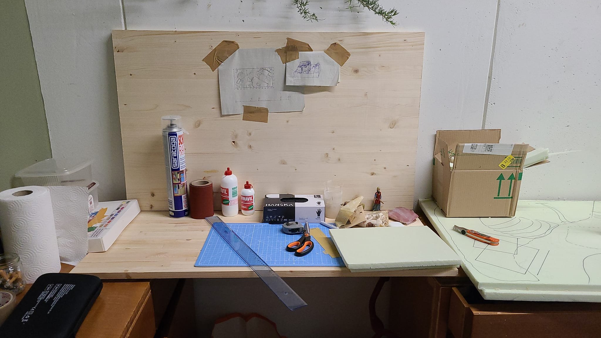 Un banco da lavoro con vari strumenti e materiali da falegnameria, tra cui forbici, squadre, colla, e scatole, con disegni e schizzi di mobili o strutture in carta appesi al pannello di legno.
