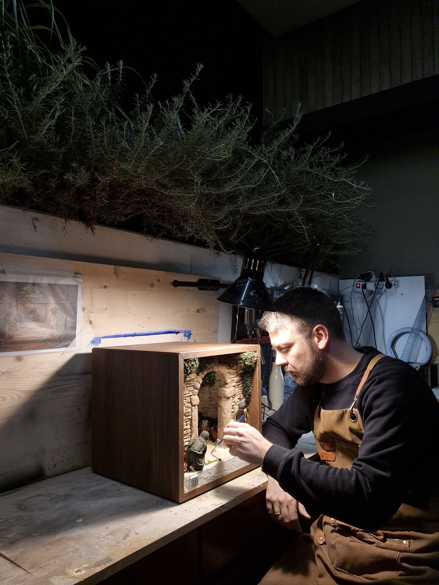 Un uomo che lavora su un modellino in miniatura di un villaggio con un arco, in un laboratorio. Sullo sfondo, una pianta e una parete in legno.