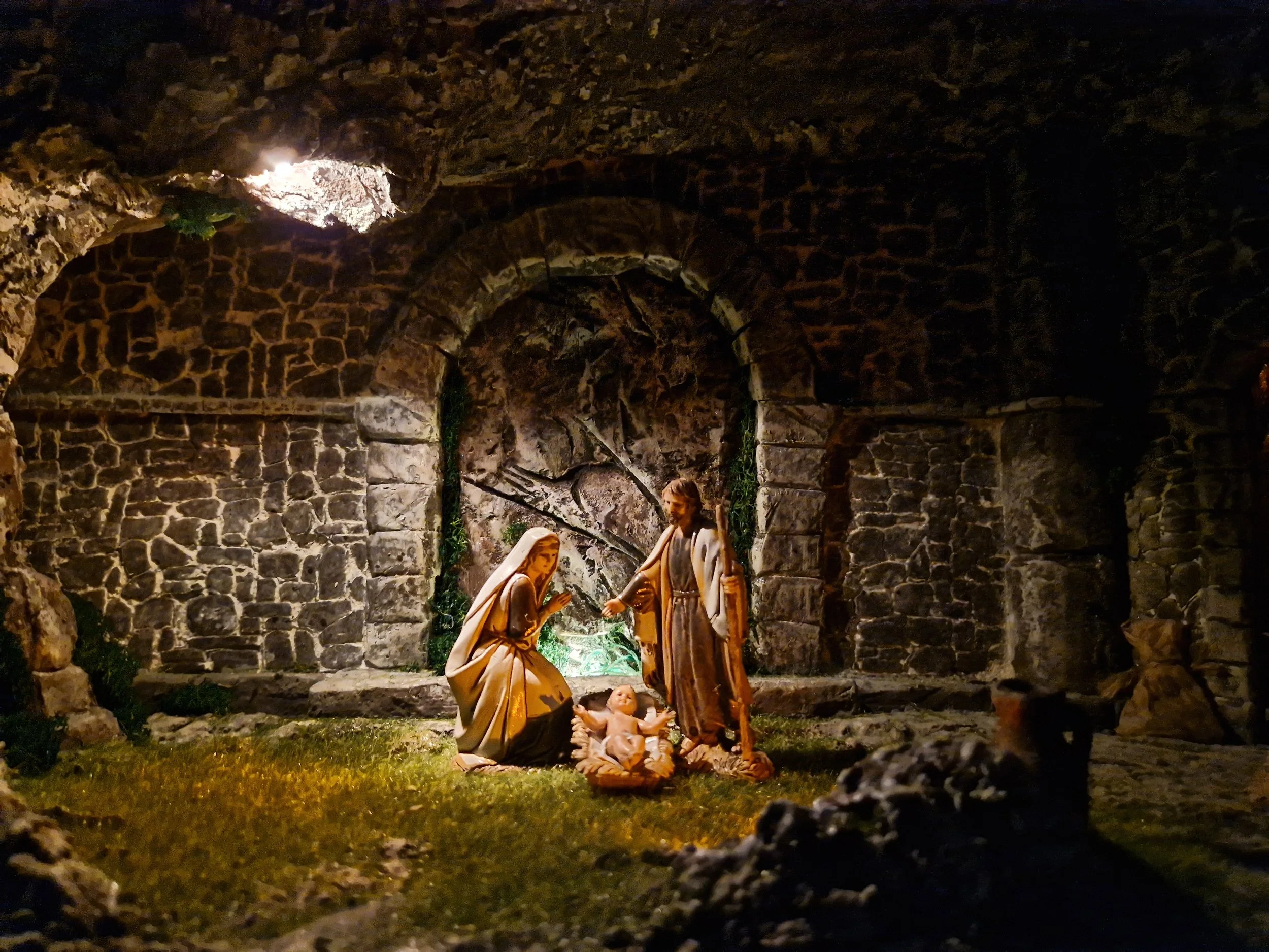 Una scena natalizia con figure di plastilina raffiguranti la Sacra Famiglia, in un ambiente che sembra una grotta di pietra, illuminata da una luce soffusa.