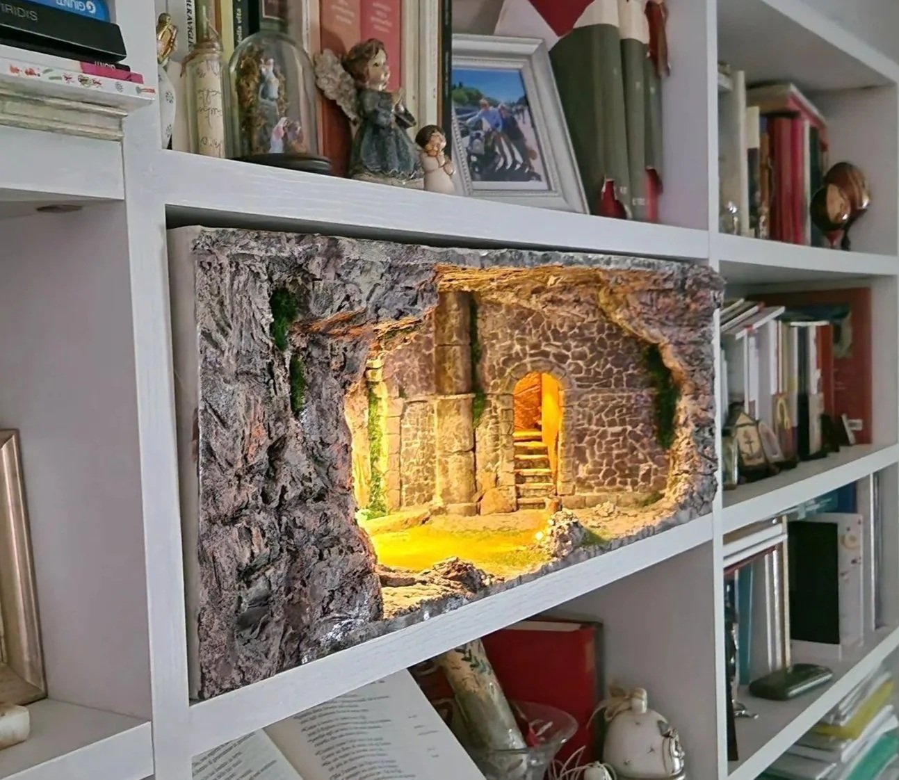 Scaffale con oggetti decorativi, libri, foto, statue e una luce calda che illumina una struttura in pietra stile grotta o rifugio