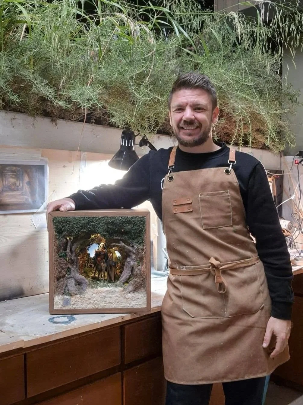Uomo con grembiule di cuoio mostra una scena natalizia in una scatola di vetro, in un laboratorio con piante sopra e una lampada.