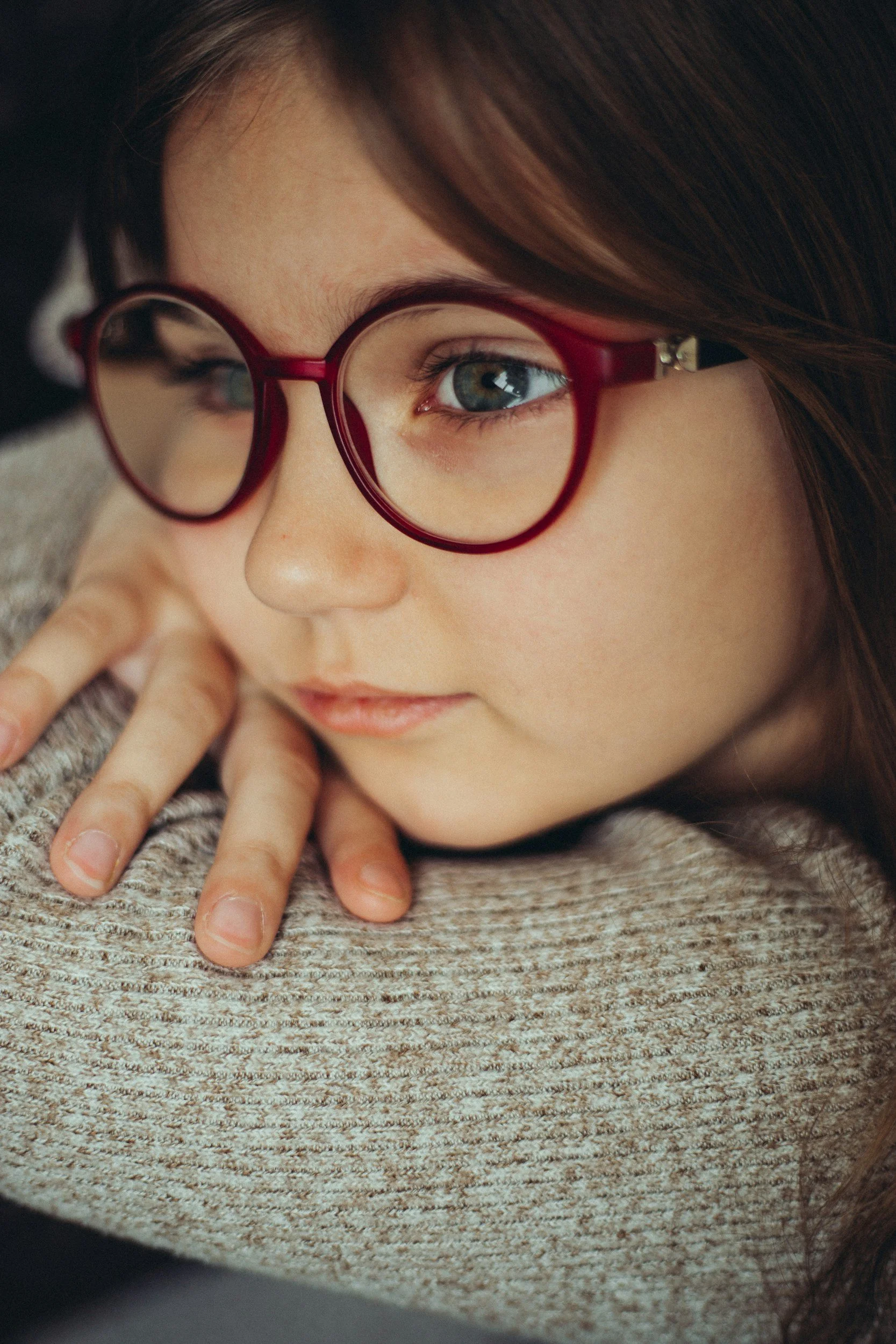 Close-up of a woman with brown hair and glasses resting her head on a textured beige surface, with her hand gently touching her face.