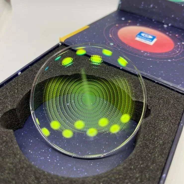A curved glass lens with concentric green rings and yellow dots, placed inside a black foam-lined box, with space-themed graphics on the box lid in the background.