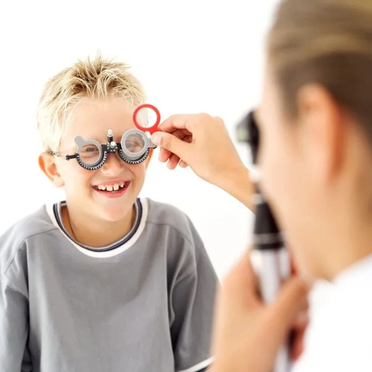 Child smiling with magnifying glasses and trial lenses during eye exam, optometrist assisting.