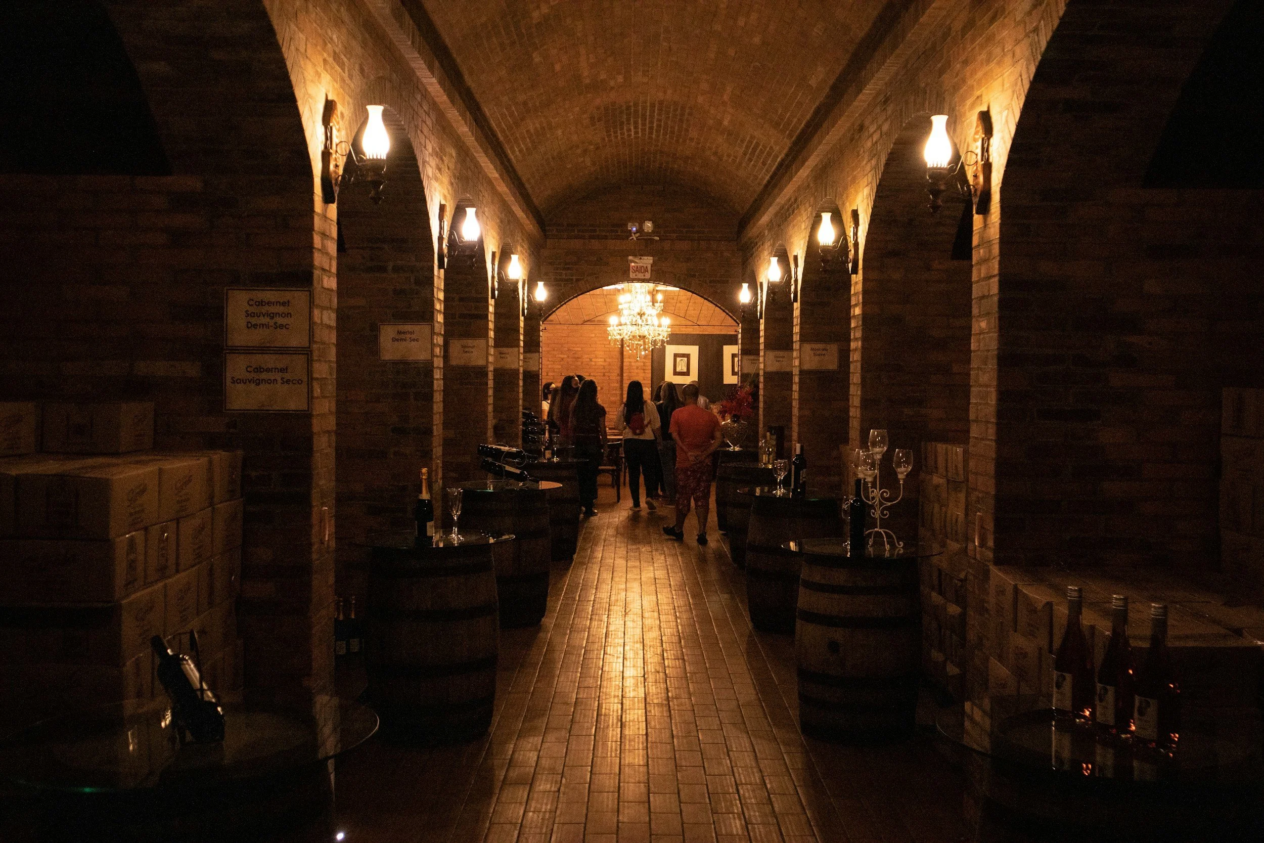 A dimly lit wine cellar with brick arches, wooden barrels, and wine bottles. People are gathered around, some looking at the bottles, with a chandelier hanging from the ceiling.