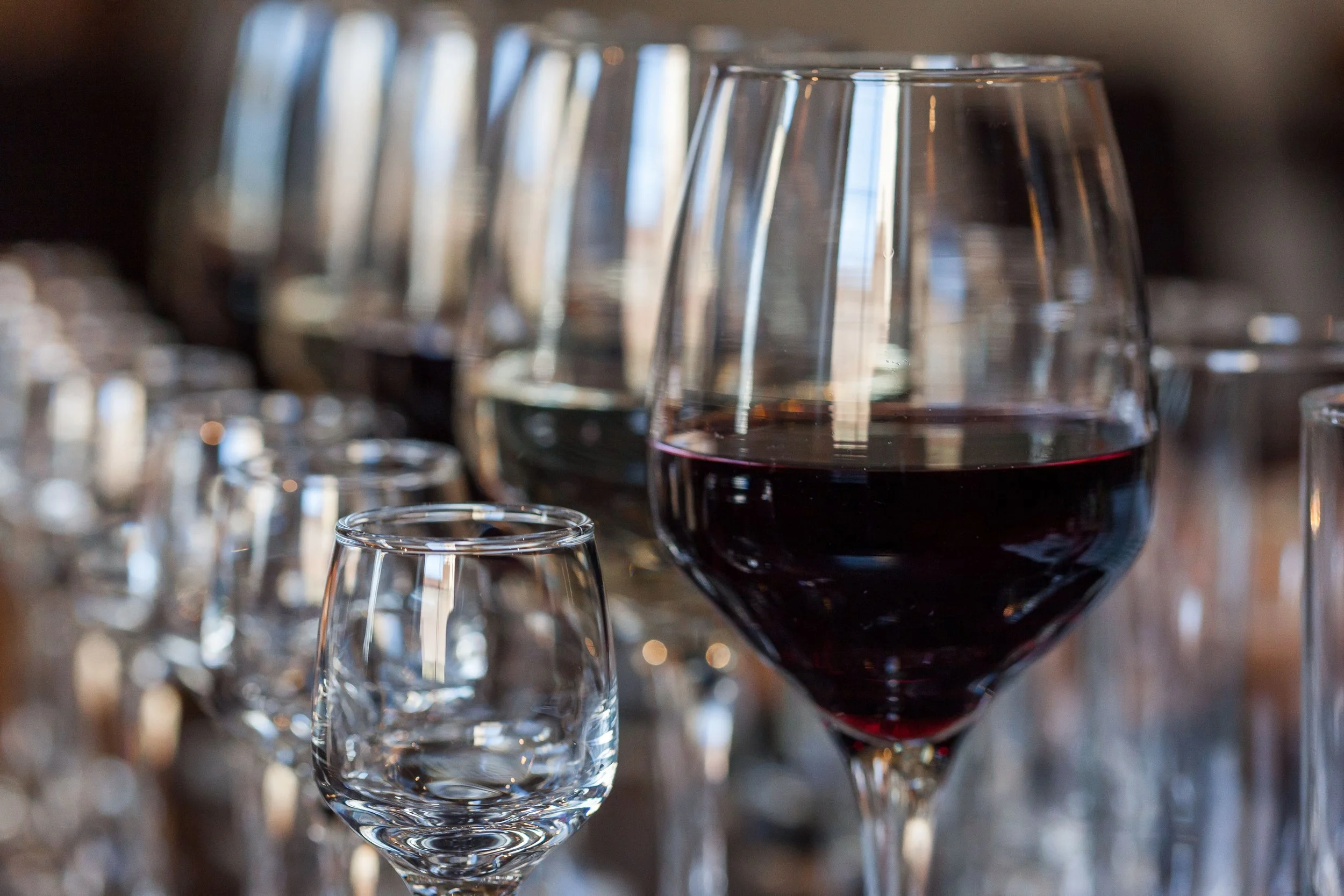A row of empty wine glasses and a filled glass of red wine on a table.