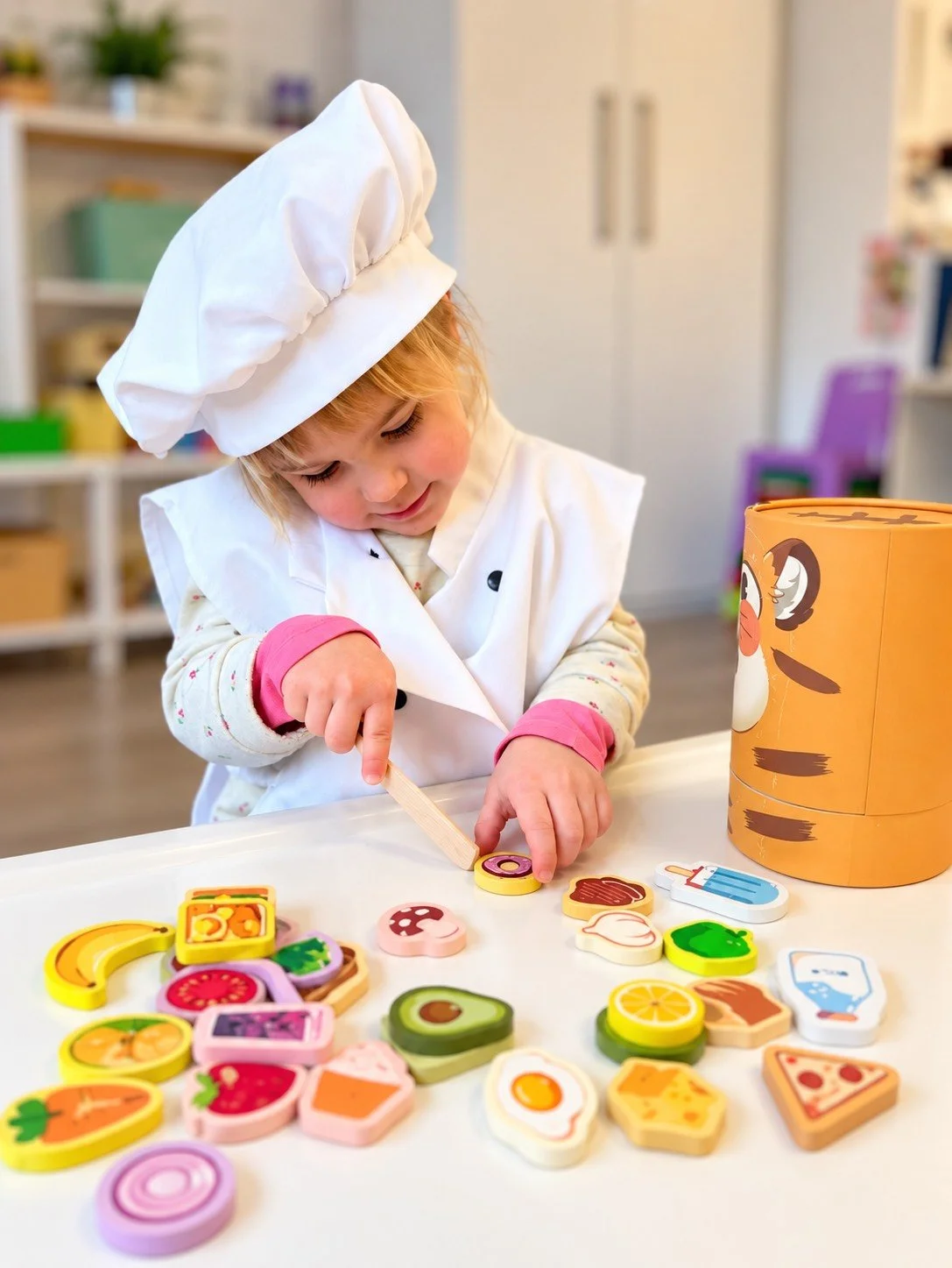 A young child dressed as a chef playing with colorful wooden toy food items on a table.