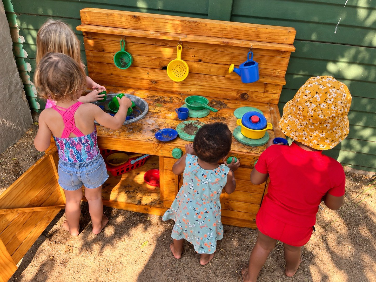 Four young children playing together at a wooden outdoor play kitchen with toy pots, pans, and utensils, some of which are dirt-stained, on a dirt ground beside a green fence.