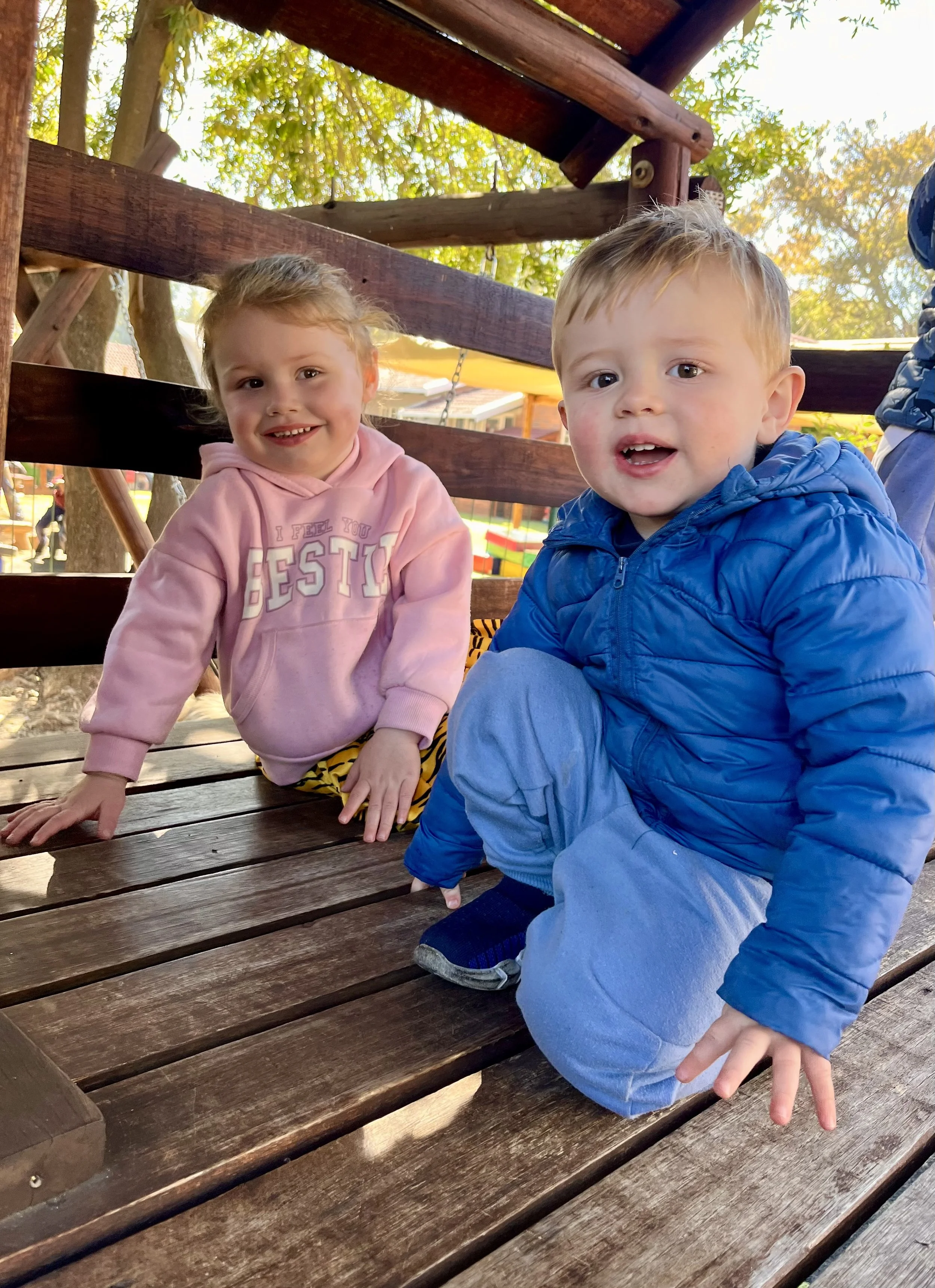Two young children, a girl and a boy, sitting on a wooden park bench outdoors, with trees and a playground in the background. The girl is wearing a pink hoodie and smiling, while the boy is wearing a blue jacket and looking at the camera.