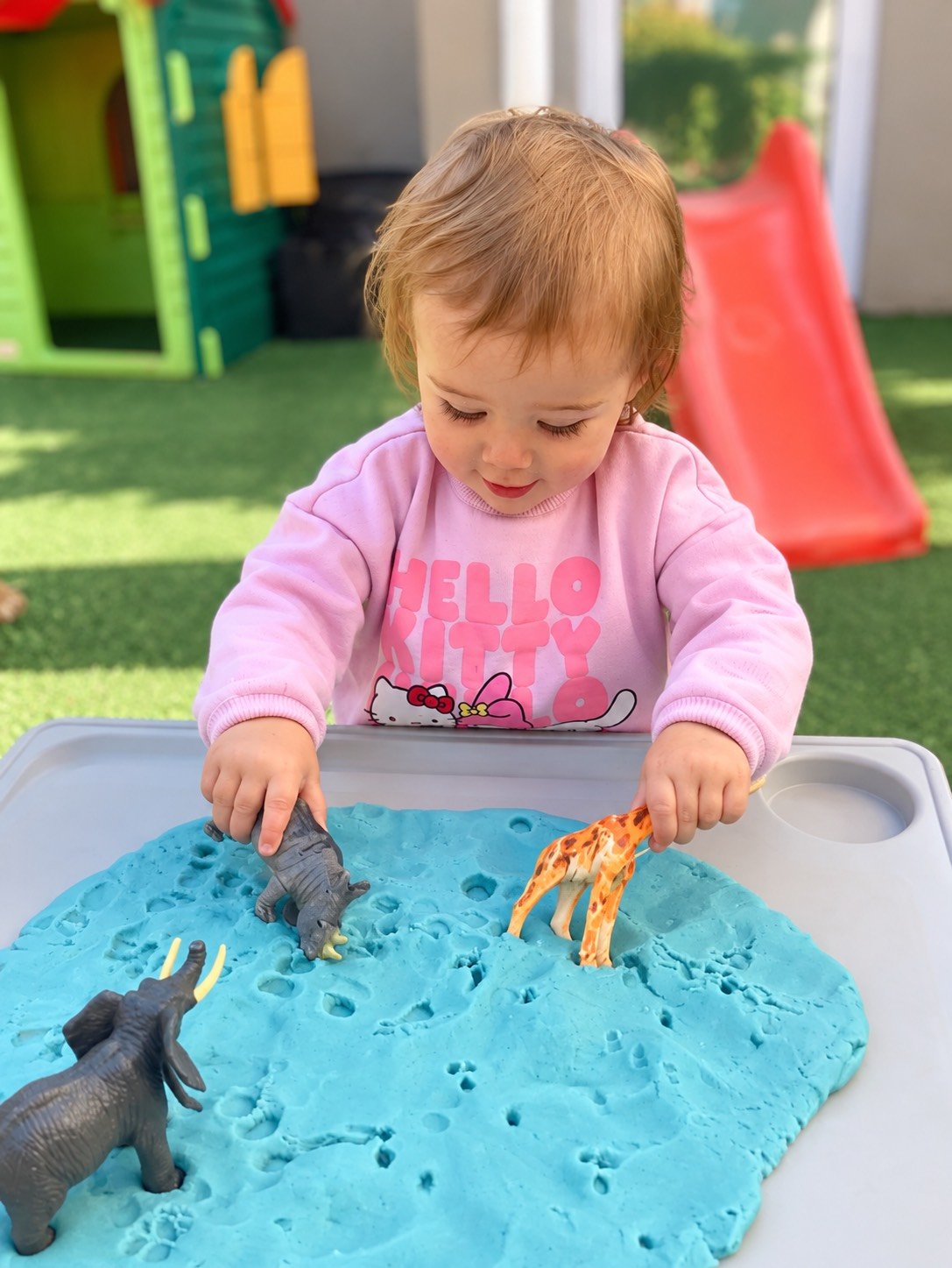 A young girl playing with toy animals in blue sand at an outdoor play area, with a red slide and colorful playhouse in the background.