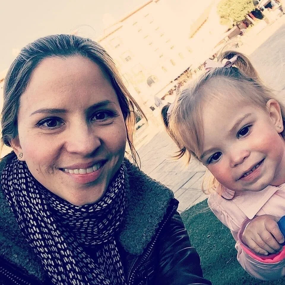 A woman taking a selfie outdoors with a young girl beside her. The woman has brown hair and is smiling, wearing a black jacket and a black-and-white scarf. The girl has blonde hair tied in pigtails with white bows, wearing a pink jacket and appears to be making a playful expression.