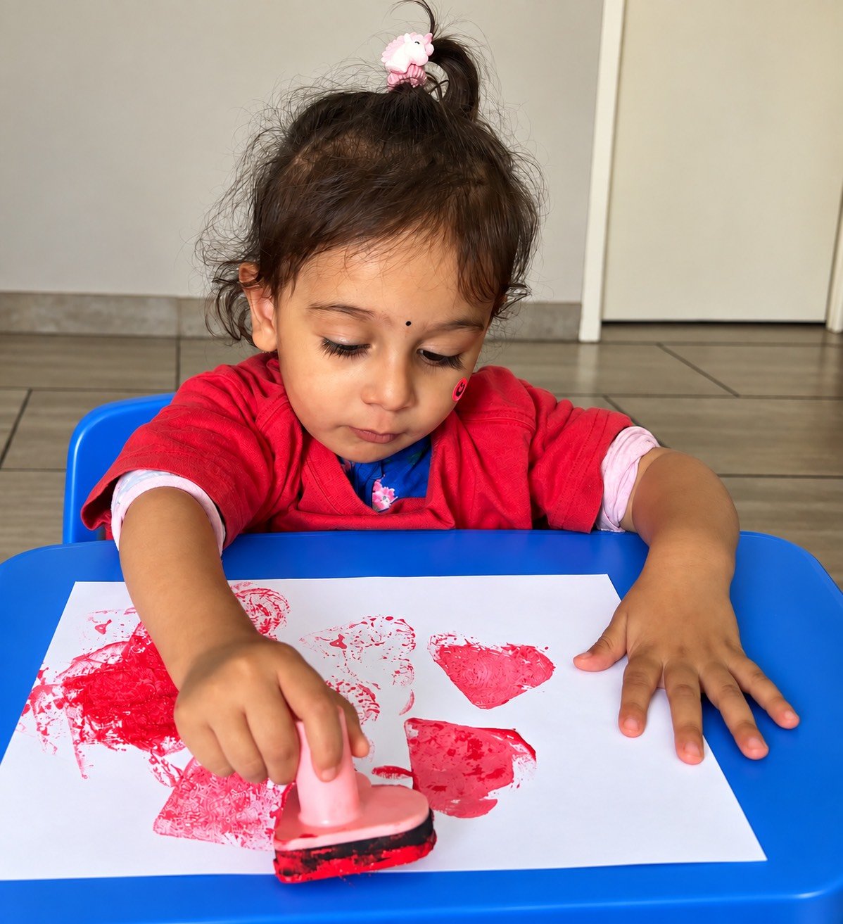 Young girl in red shirt stamping red paint onto a white sheet of paper, with heart shapes, at a blue table.