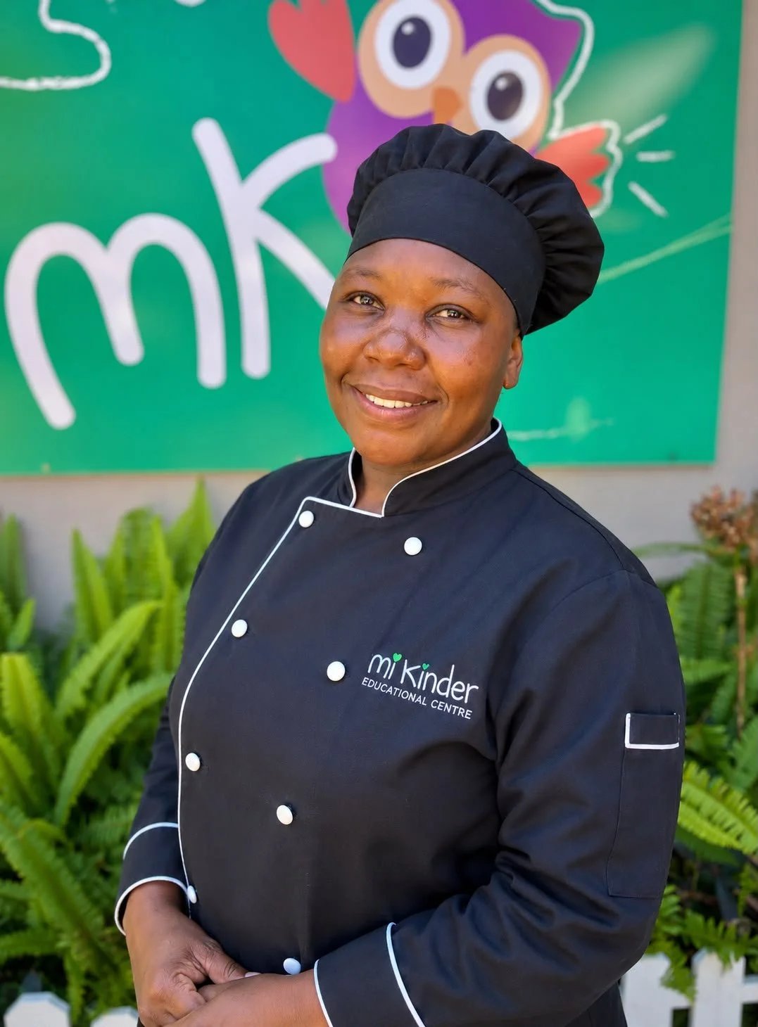 A smiling female chef wearing a black chef's uniform and hat, standing outdoors in front of a colorful background with the logo 'mK' and some vibrant green plants.