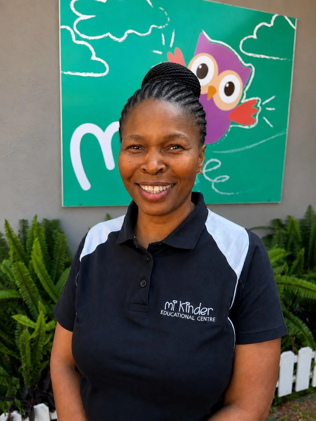 Smiling woman standing outdoors in front of a colorful owl-themed mural with bright green background, wearing a black polo shirt with white accents and the logo 'Mi Kinder Educational Centre'.