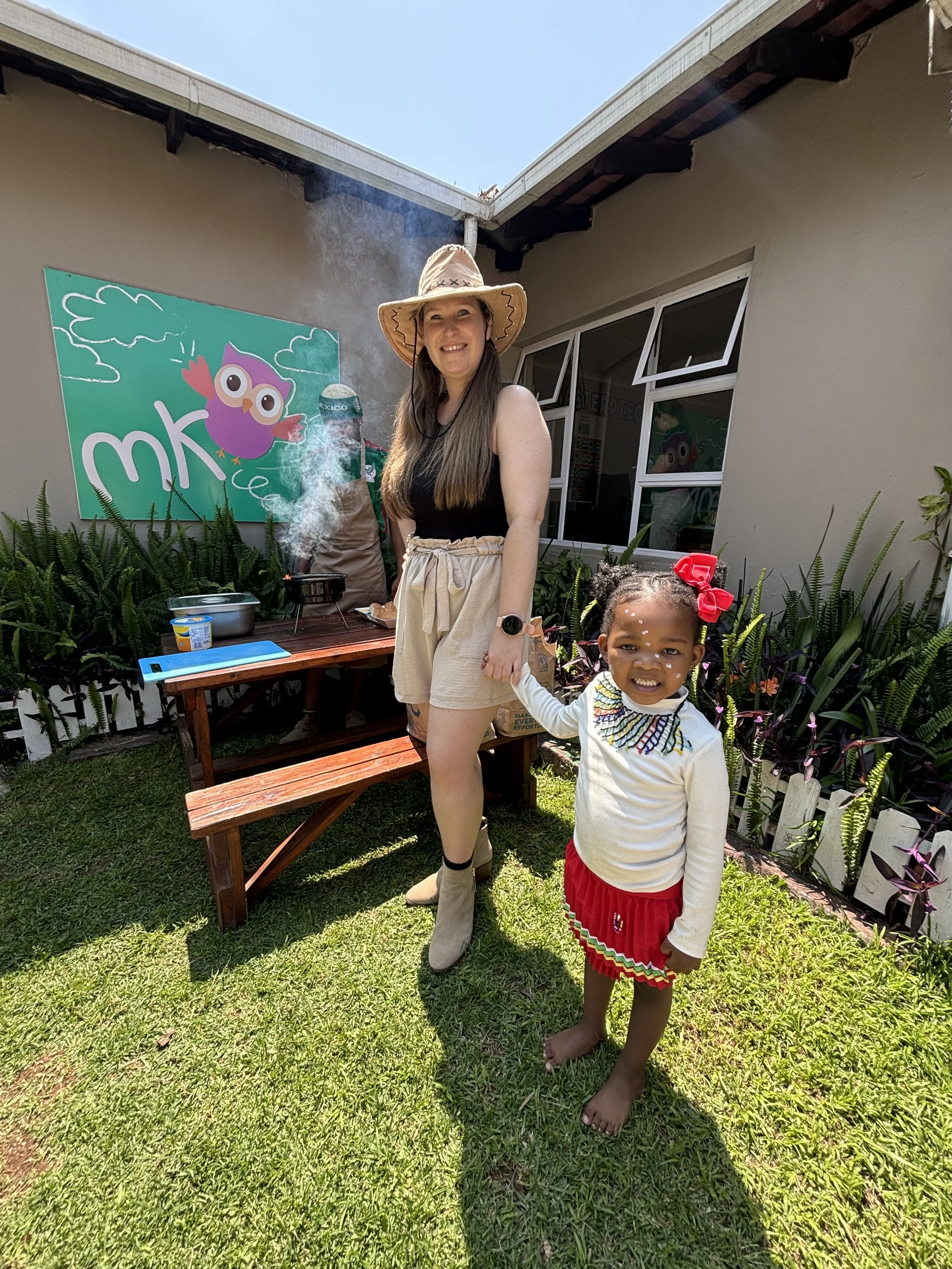 A woman and a young girl stand outside in a garden on a sunny day. The woman is wearing a wide-brimmed hat, black top, and beige shorts, smiling at the camera. The girl, holding the woman's hand, is barefoot, with face paint, wearing a white sweater 