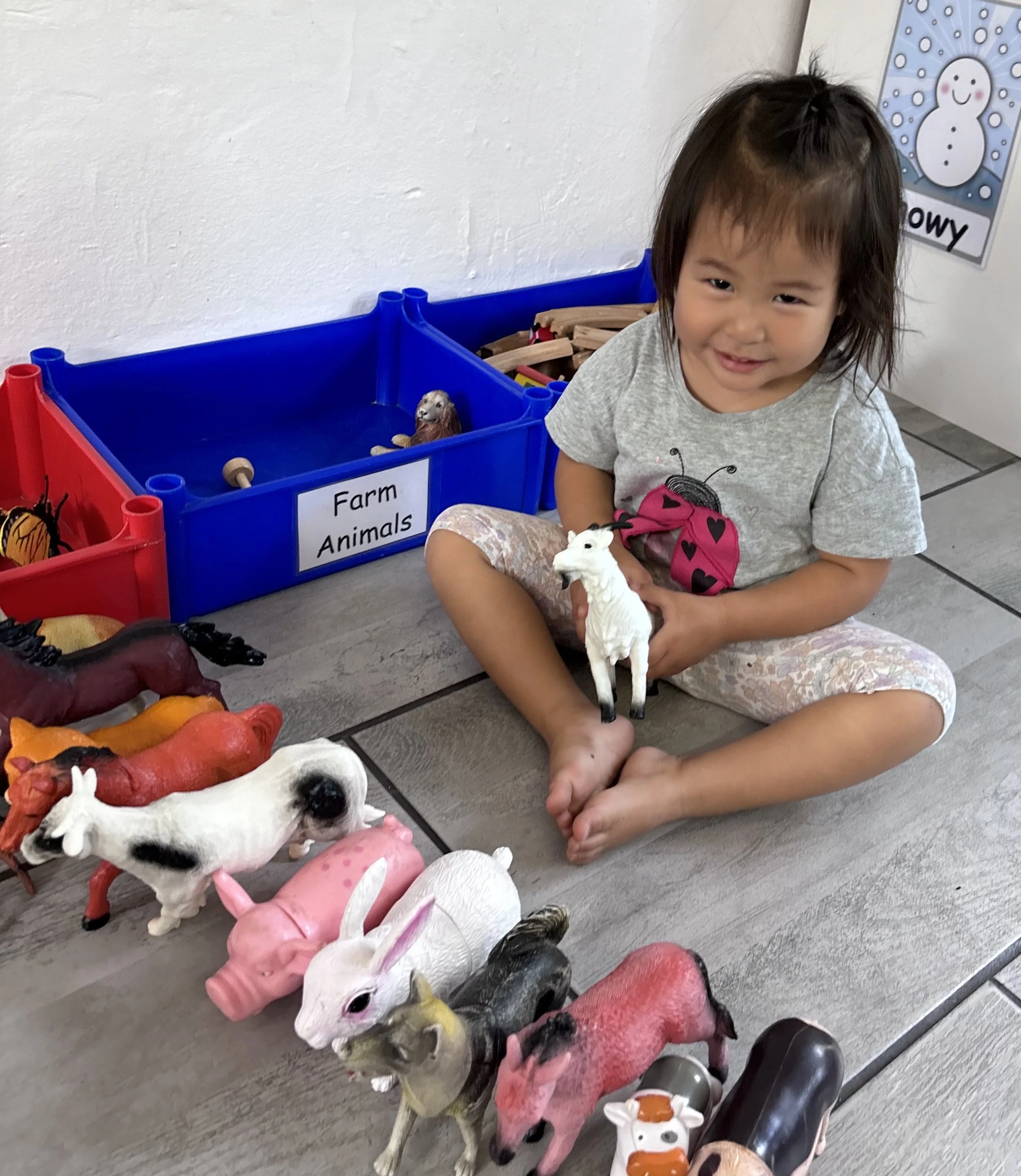A young girl with brown hair and a gray shirt sitting on the floor, smiling, holding a white toy horse. In front of her, there are various toy farm animals including a cow, pig, horse, duck, and chicken, arranged on the floor. Behind her, a blue stor