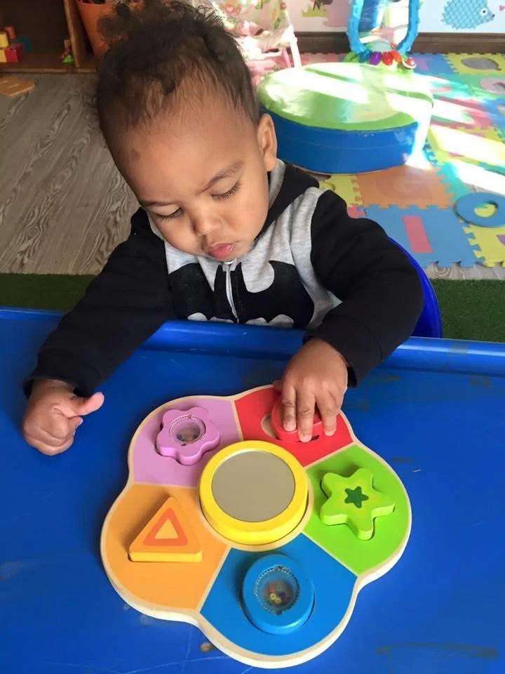 A young child playing with a colorful shape-sorting toy at a blue table, in a playroom with foam puzzle mats and toys in the background.