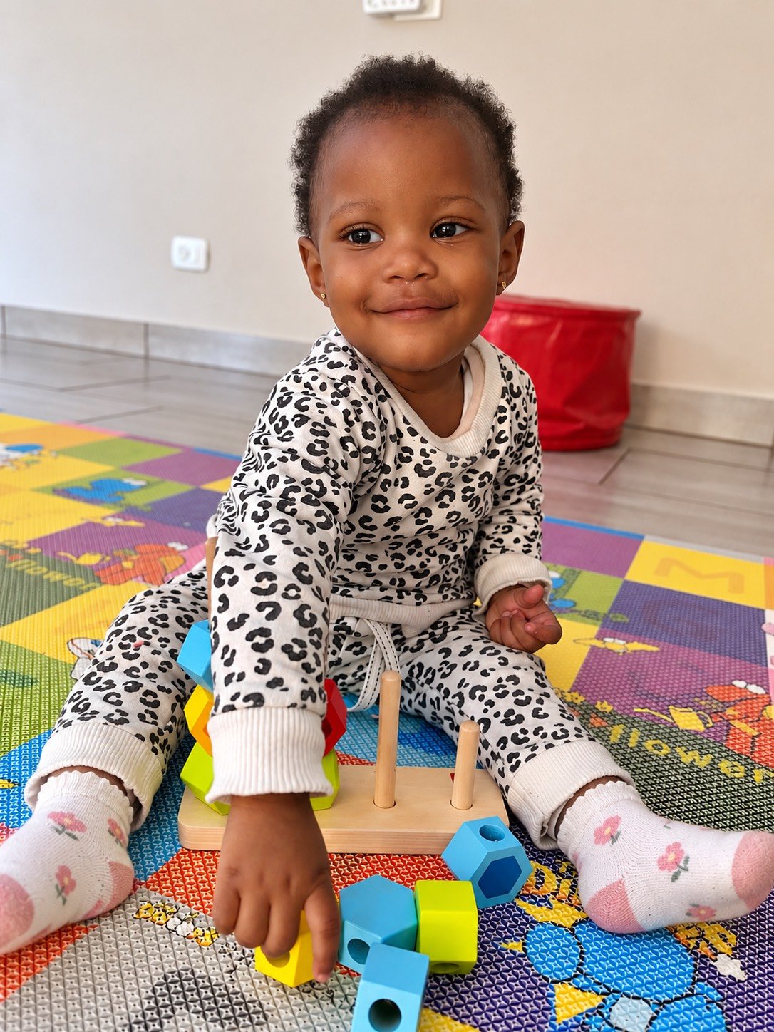 A young girl with curly hair and earrings, wearing a white and black leopard-print outfit, sitting on a colorful foam playmat, playing with wooden and plastic toys, and smiling at the camera.