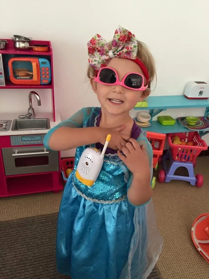 A young girl with a large floral bow and pink sunglasses is dressed as a princess and smiling while holding a toy microphone in a playroom with colorful kitchen toys and shopping cart.